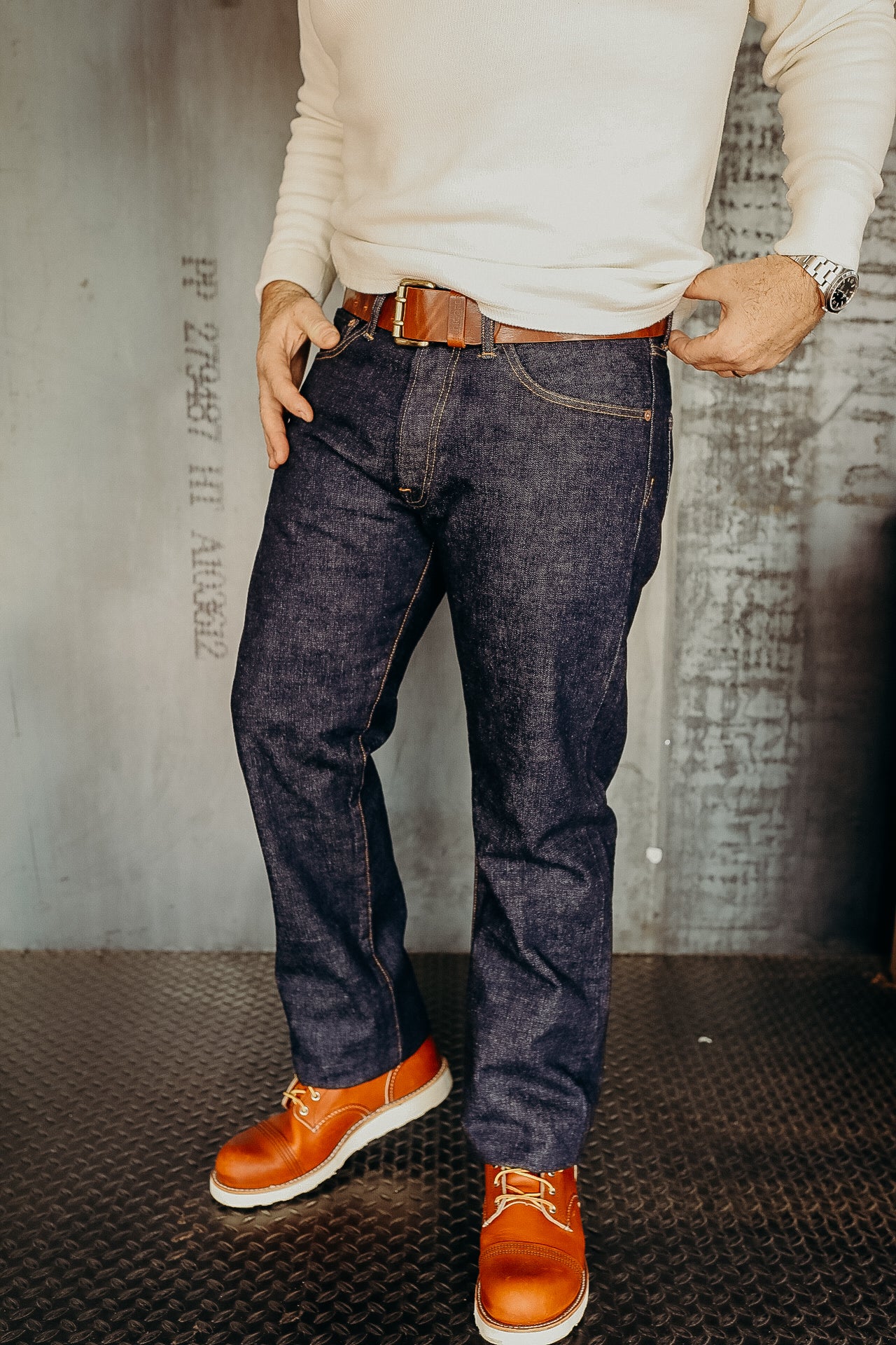 Man wearing dark blue denim jeans, brown leather belt, white long-sleeve shirt, and tan leather boots indoors
