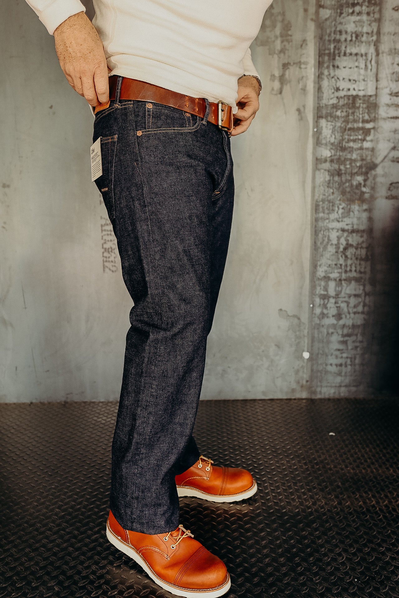 Man wearing dark raw denim jeans with brown leather belt and orange leather work boots on metal flooring