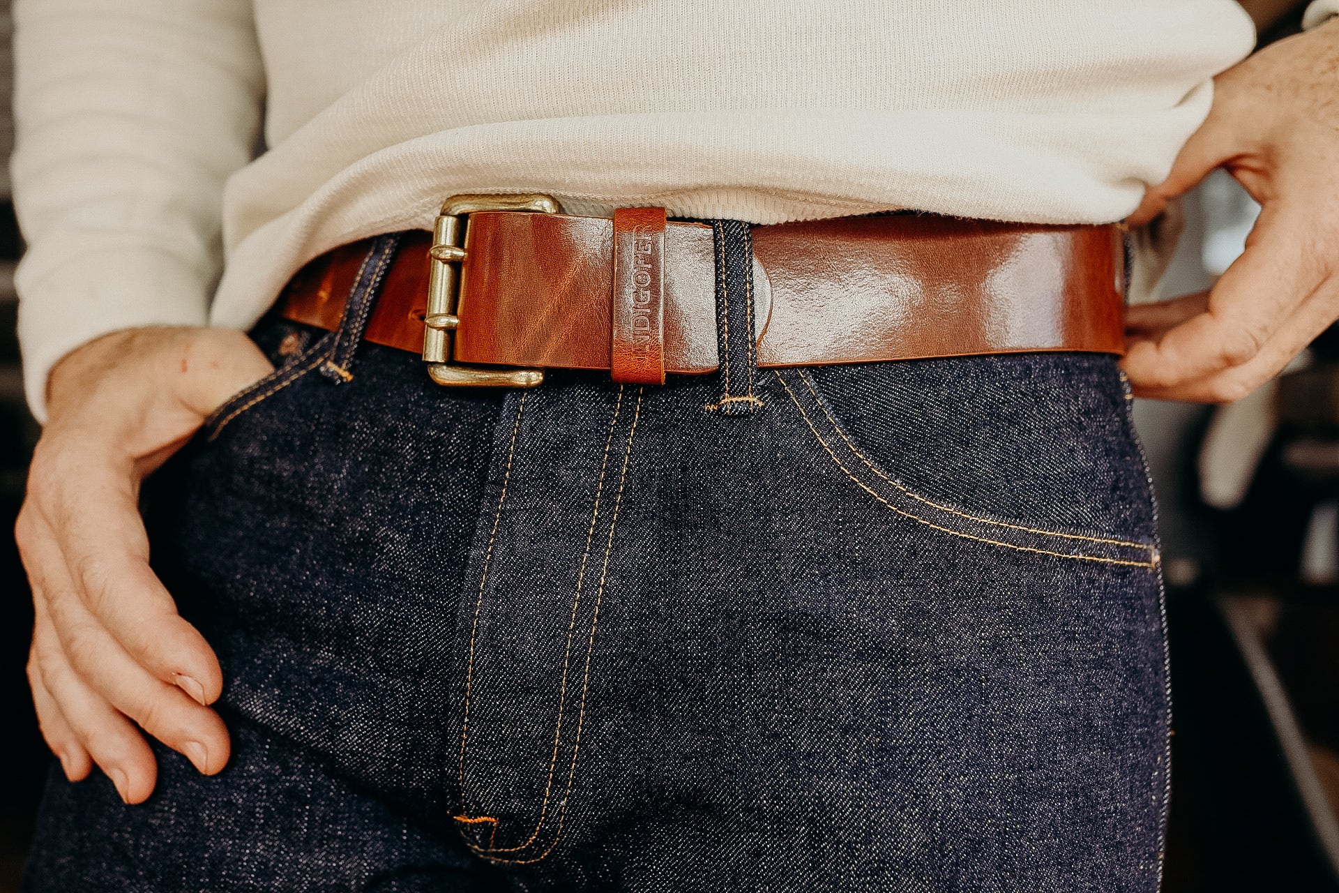 Close-up of a person wearing dark denim jeans with contrast stitching and a polished brown leather belt with brass buckle