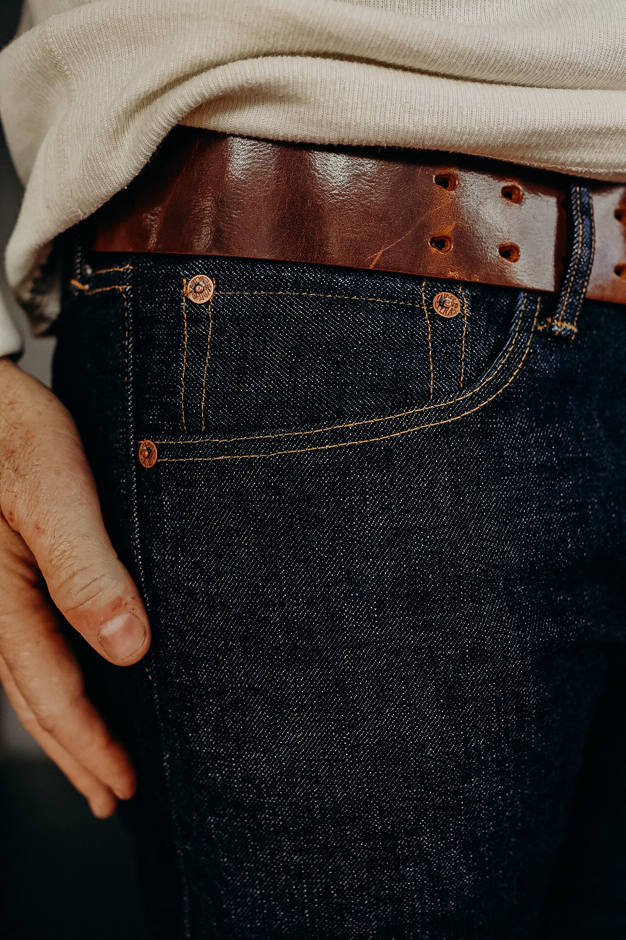 close-up of dark denim jeans with copper rivets paired with a brown leather belt and cream sweater