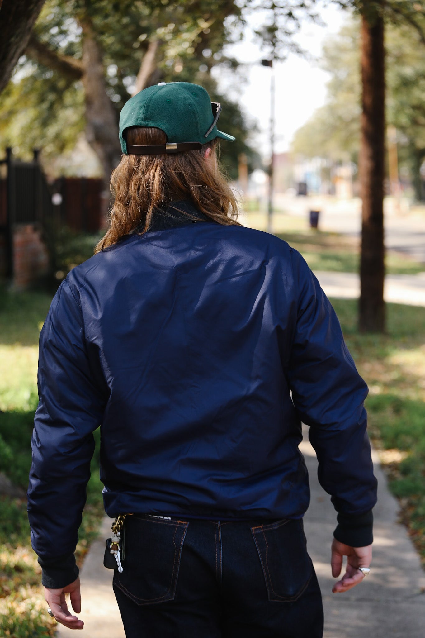 Person wearing a blue jacket and green cap walking on a sidewalk with trees and grass in the background