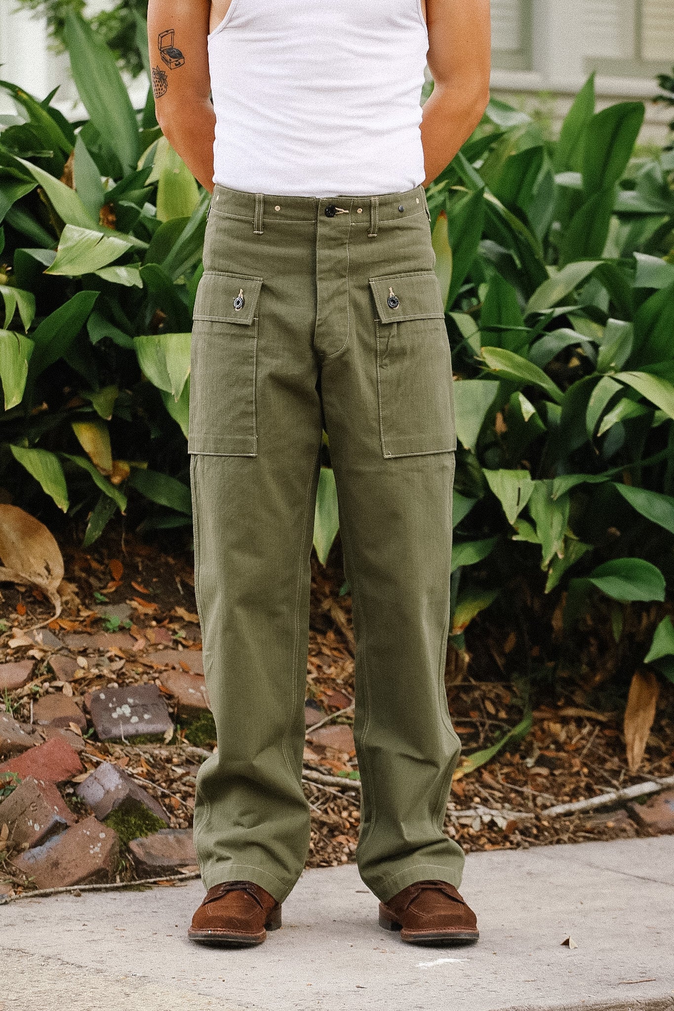 Person wearing green cargo pants and a white tank top standing outdoors with plants in the background.