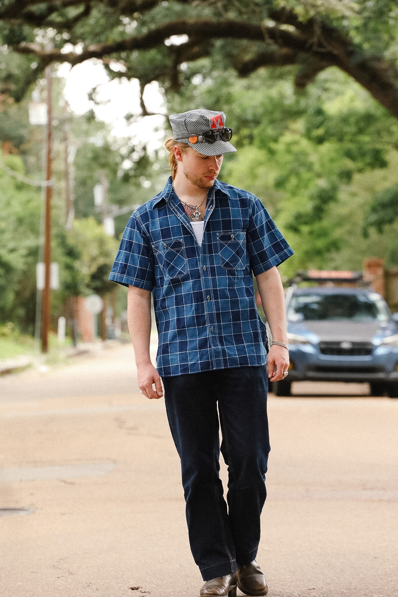 Man wearing a blue plaid shirt and cap walking on a street with trees and cars in the background.