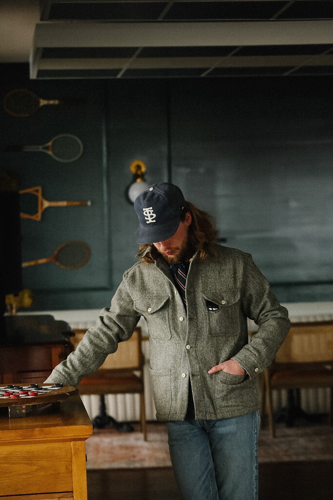 Man in a green jacket and cap standing in a room with sports equipment on the wall.