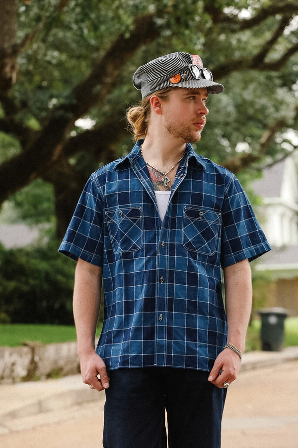 Man wearing a blue plaid shirt and cap outdoors with trees in the background