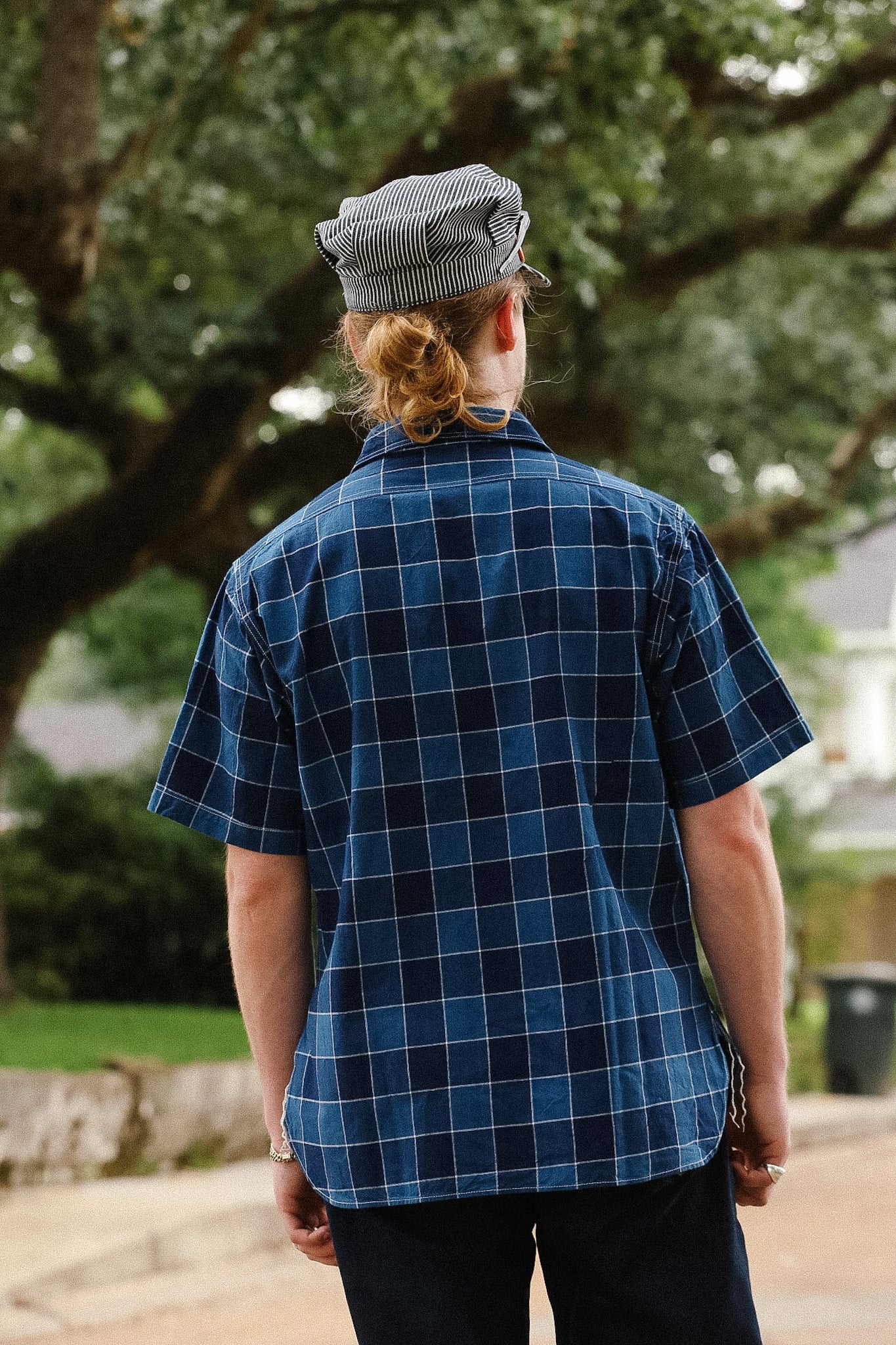 Person wearing a blue plaid shirt and cap standing outdoors with trees in the background