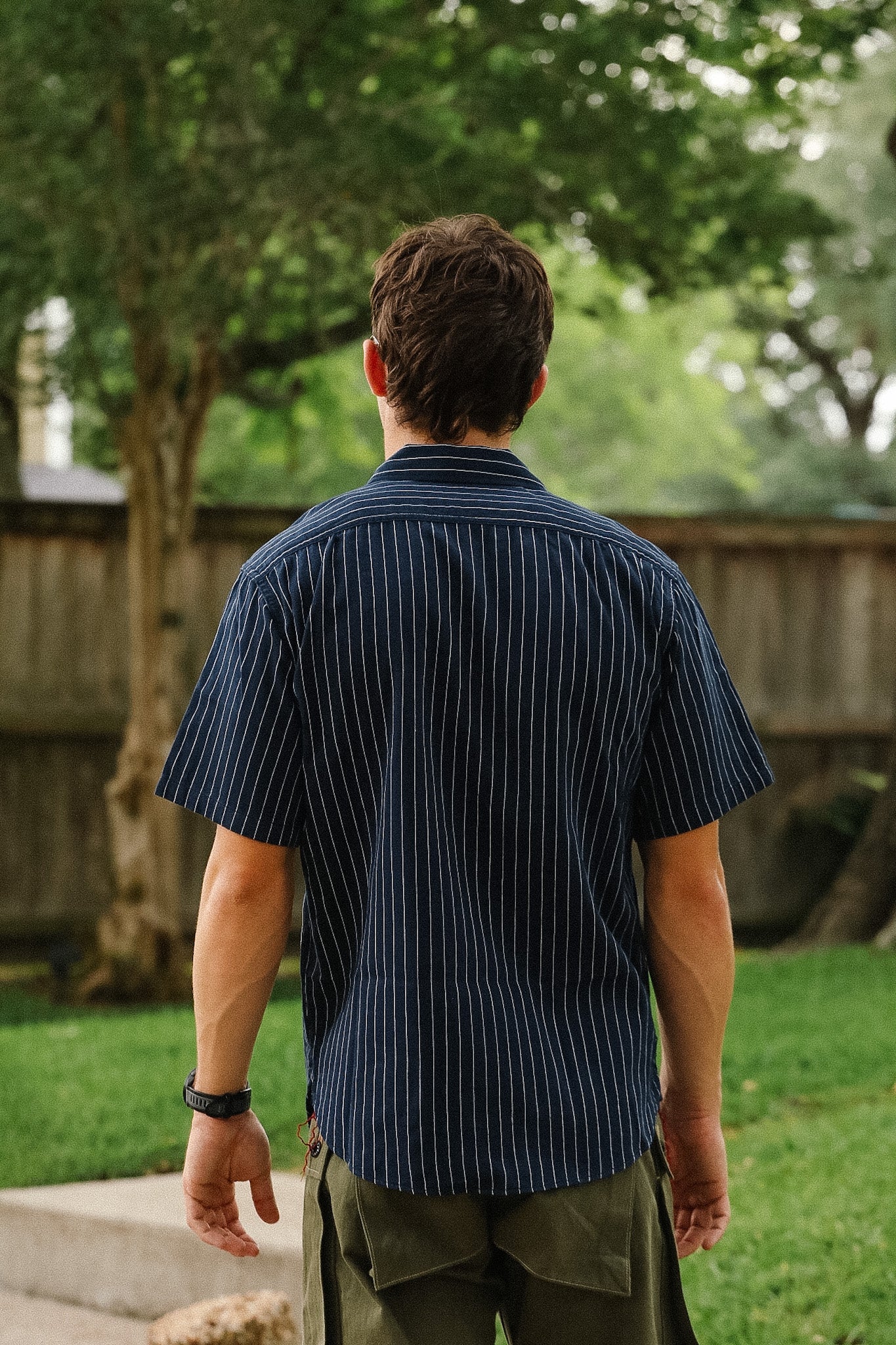 Man wearing a dark blue striped shirt standing outdoors with trees and grass in the background