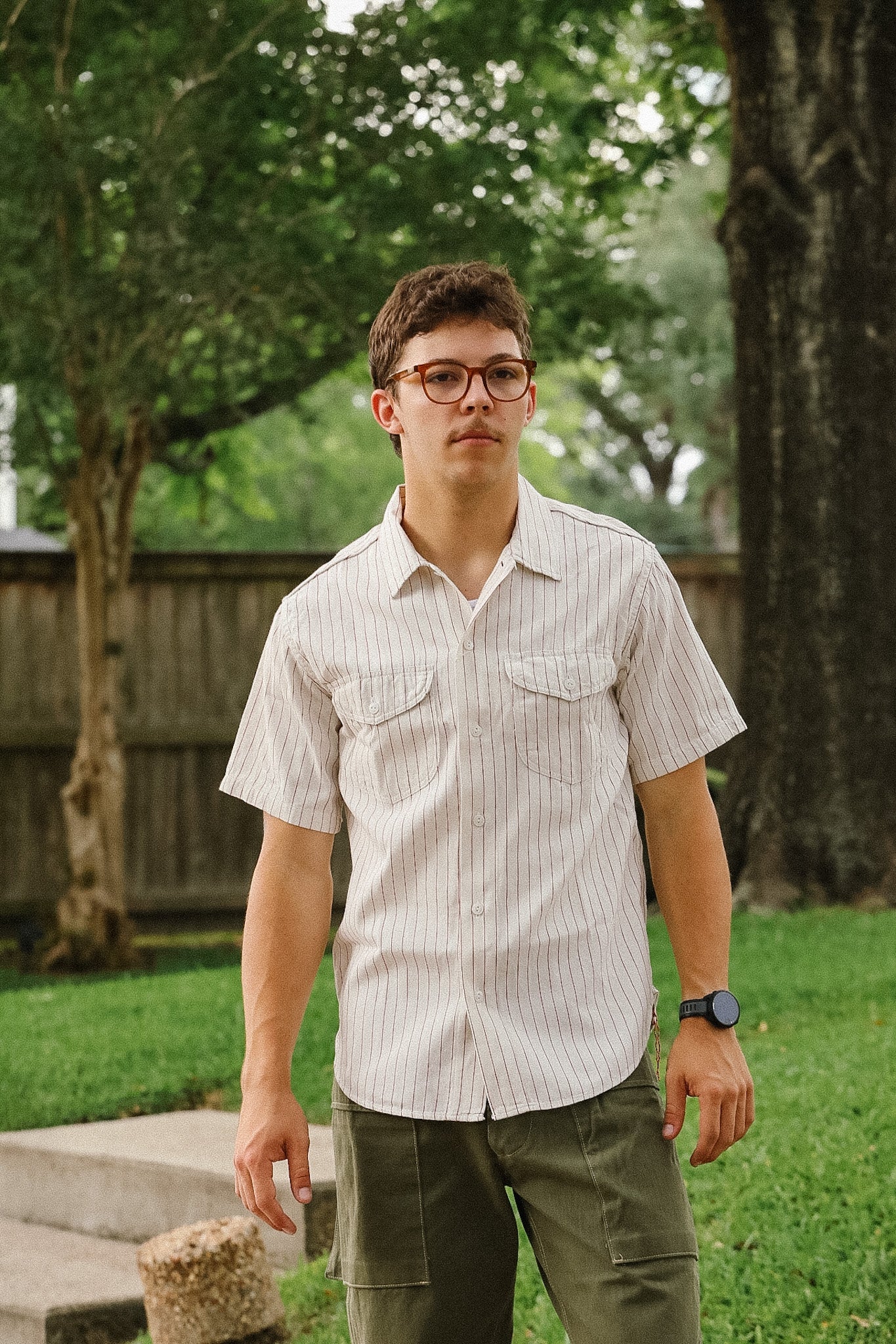 Man wearing a striped shirt and glasses standing outdoors with trees and grass in the background