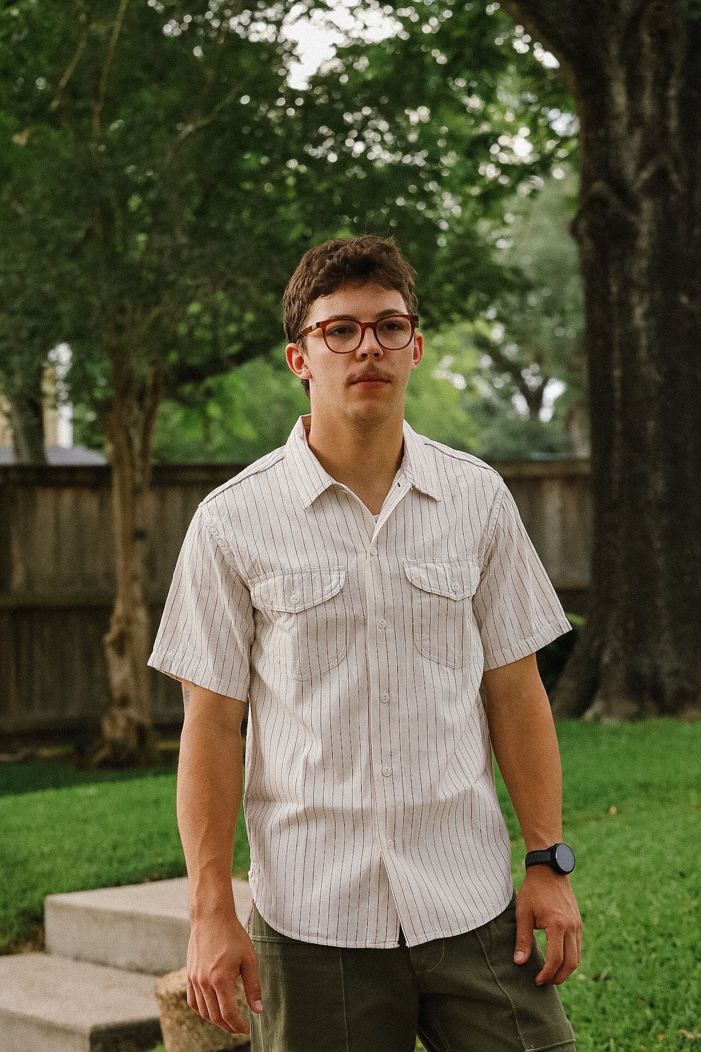 Man wearing a light-colored striped shirt and glasses standing outdoors with trees in the background