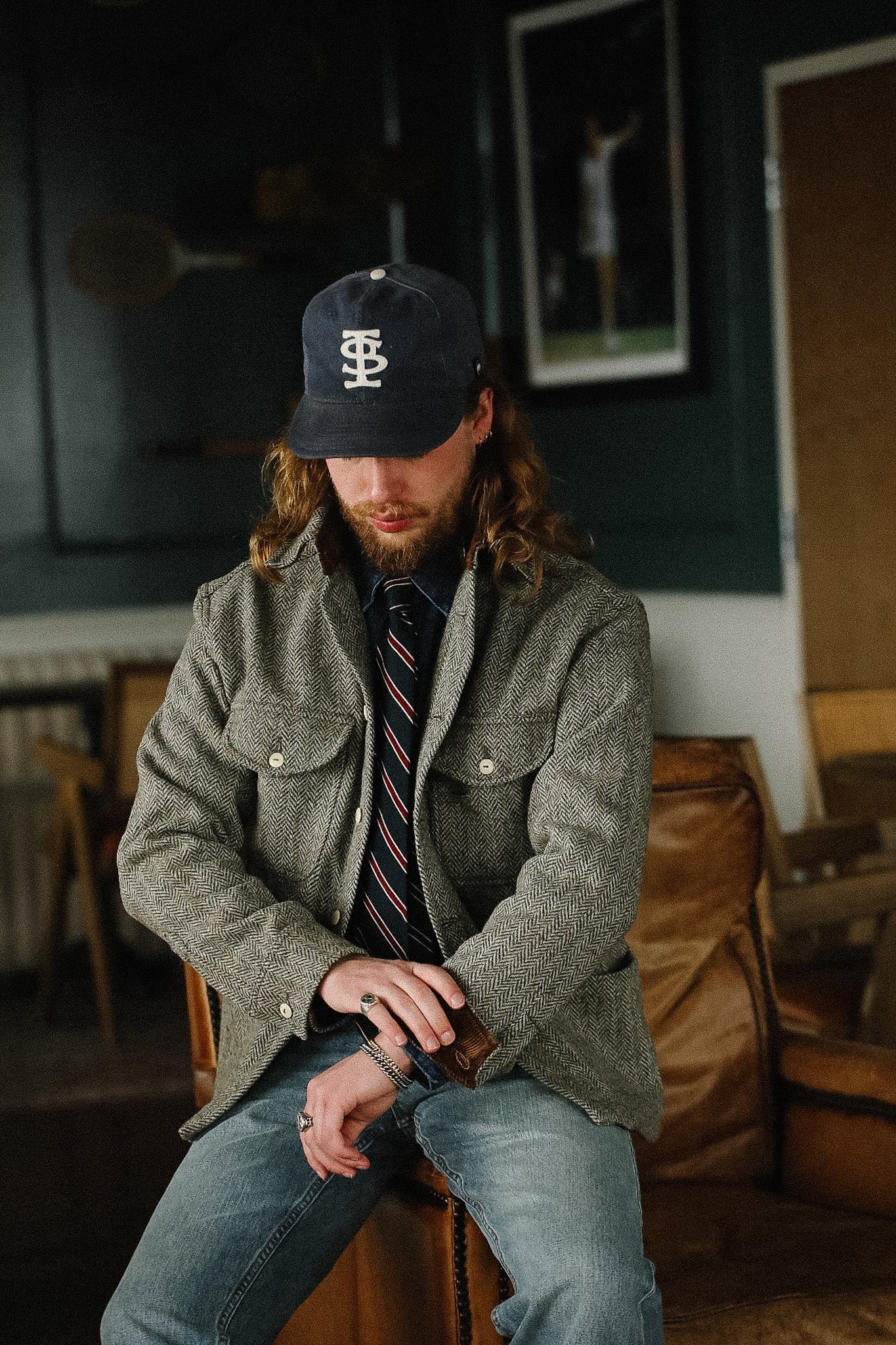 Man wearing a cap and coat sitting in a room with a deer painting on the wall.
