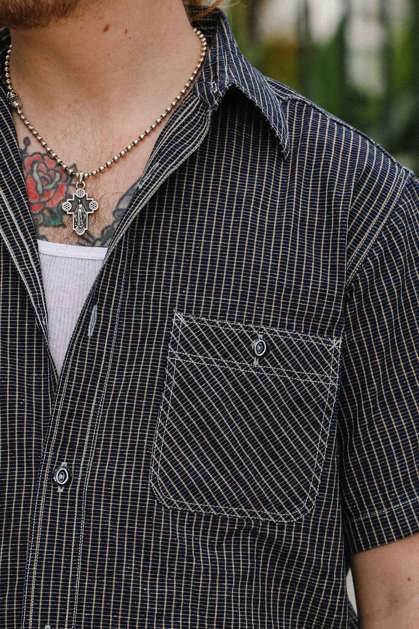 Person wearing a black checkered shirt with a pocket, visible tattoos, and a necklace.