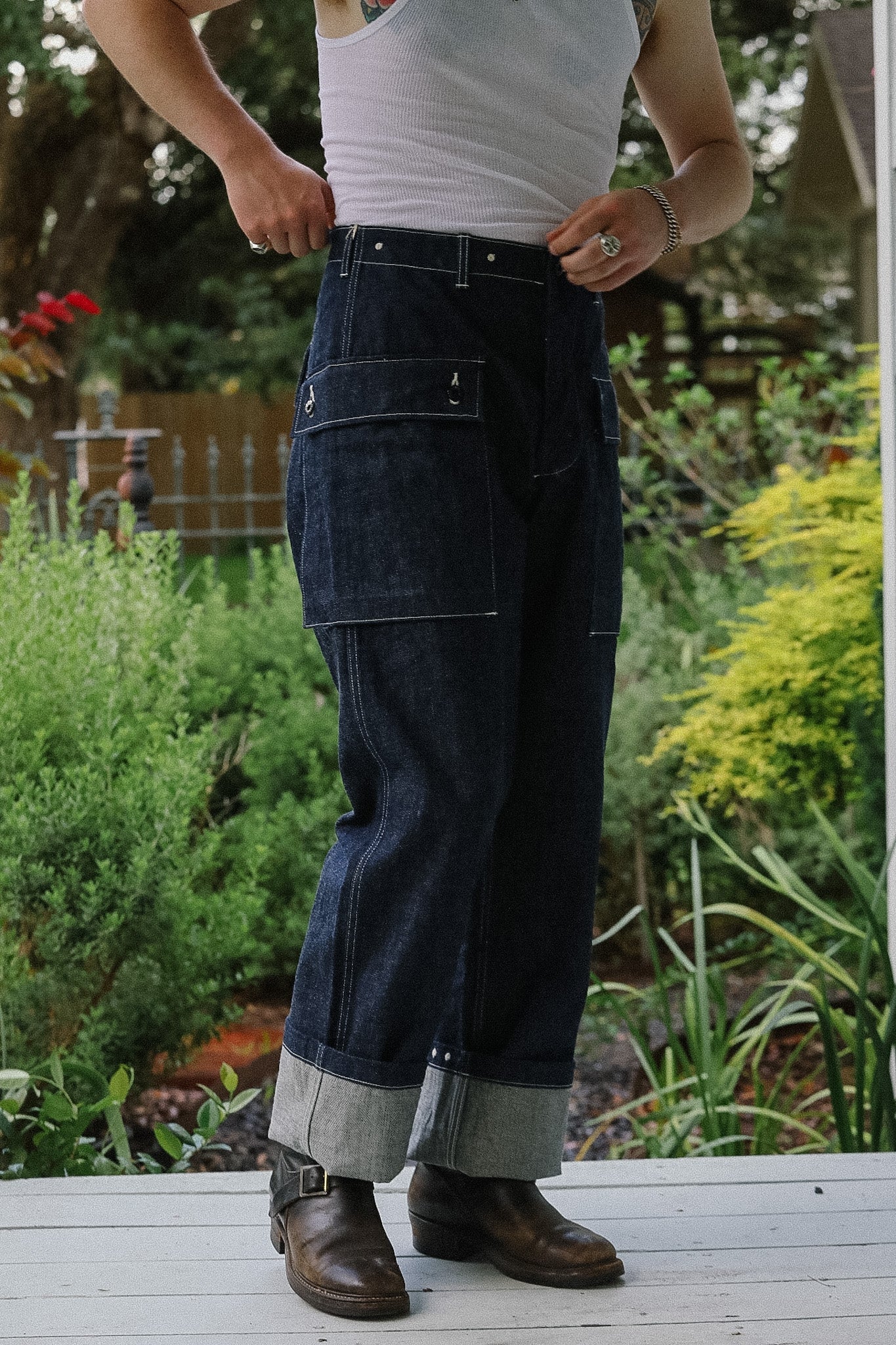 Person wearing a white tank top and dark blue cargo pants standing on a wooden deck with greenery in the background.