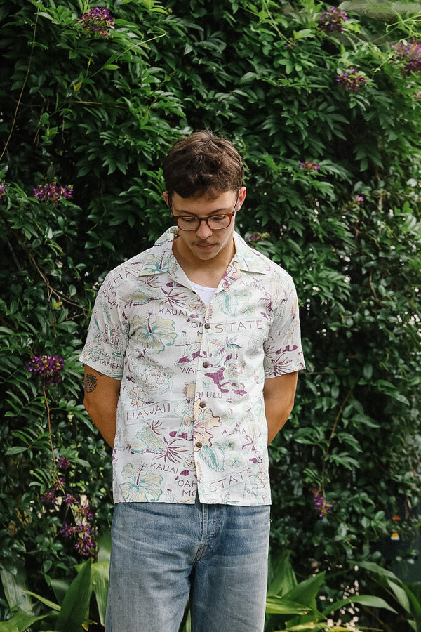 Man wearing a patterned shirt and glasses standing in front of green foliage.