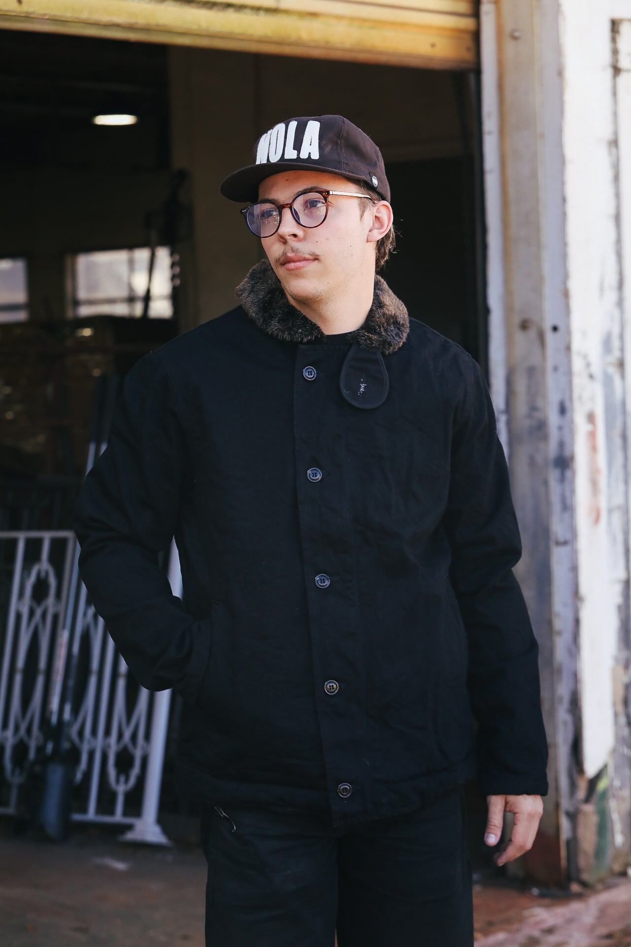 Person wearing a black coat and cap with 'HOLA' text, standing in an urban setting.