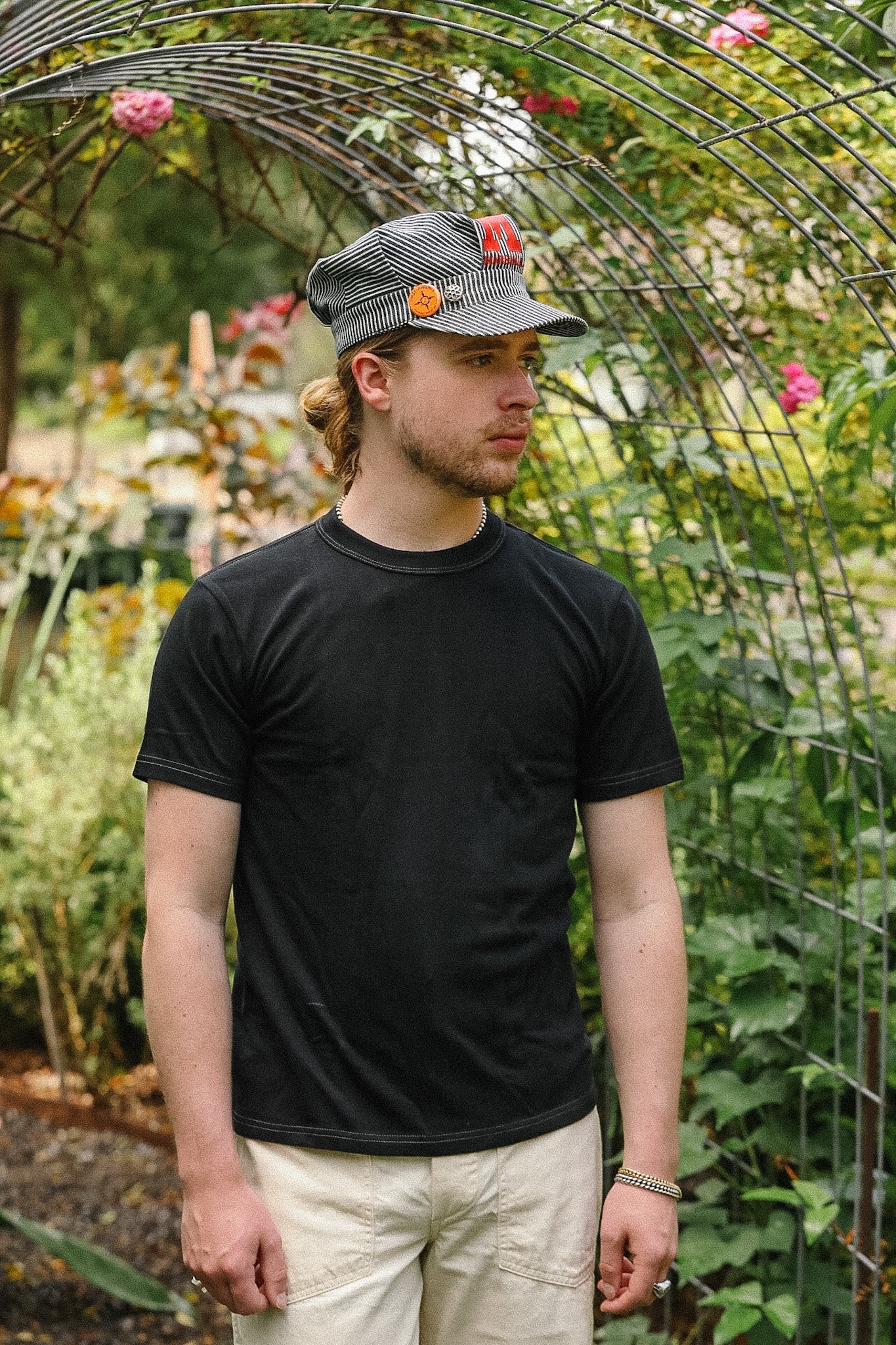 Man wearing a black t-shirt and cap standing in a garden with plants and flowers in the background