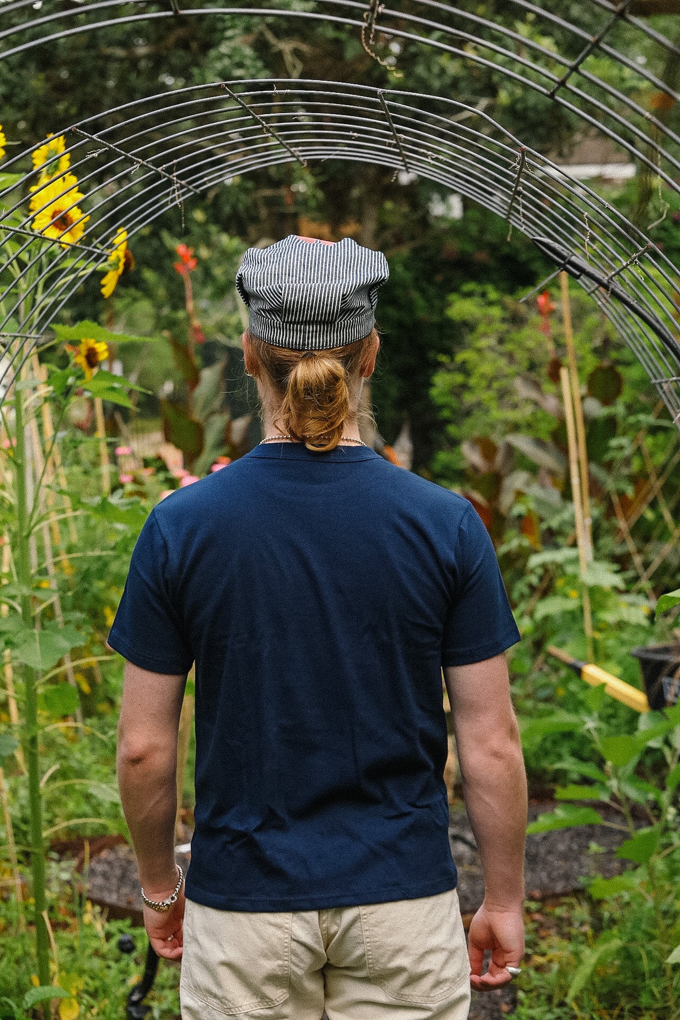 Person wearing a blue shirt and cap walking through a garden with plants and sunflowers.