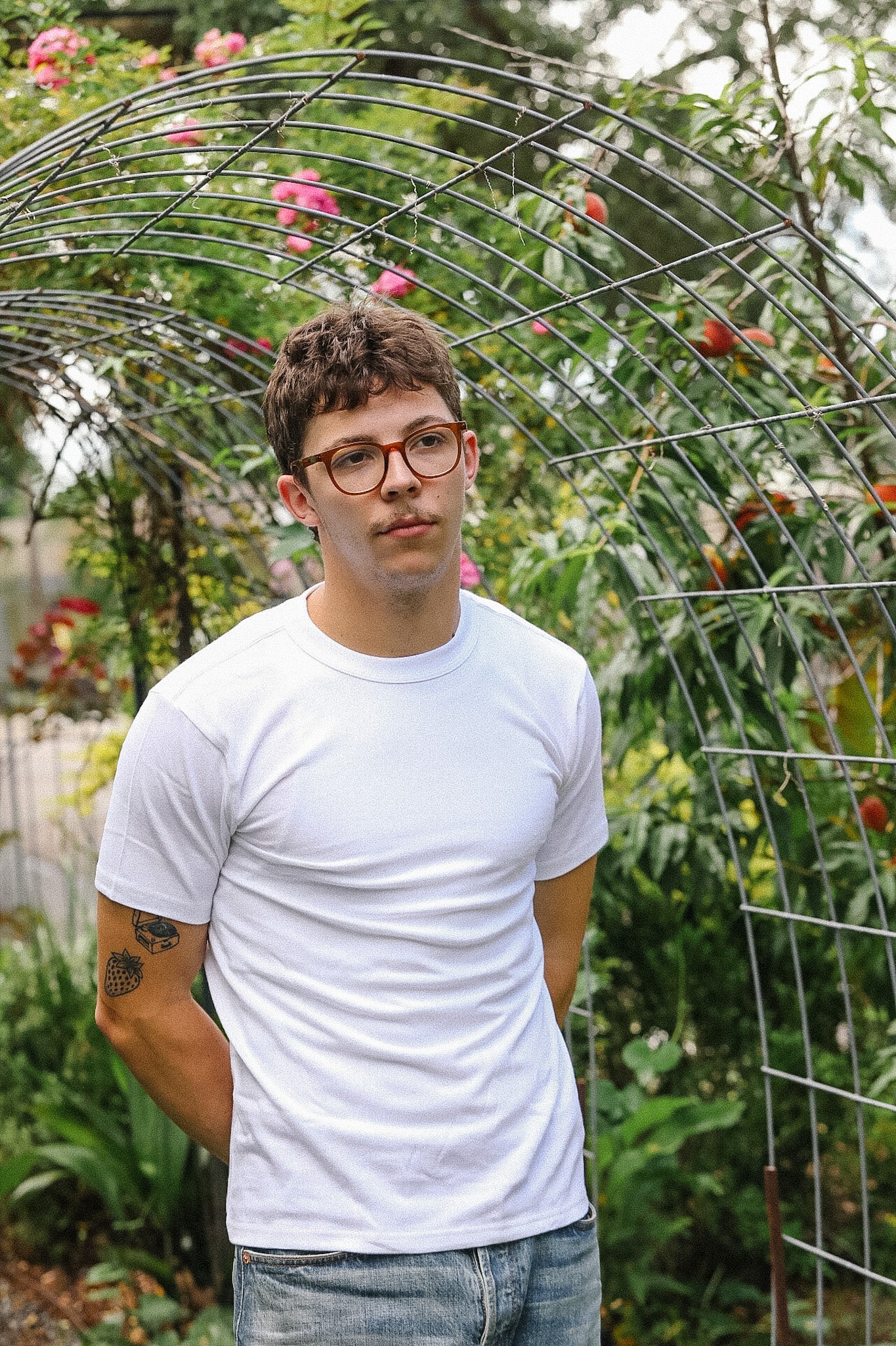 Man wearing glasses and a white t-shirt standing in front of a metal archway with greenery