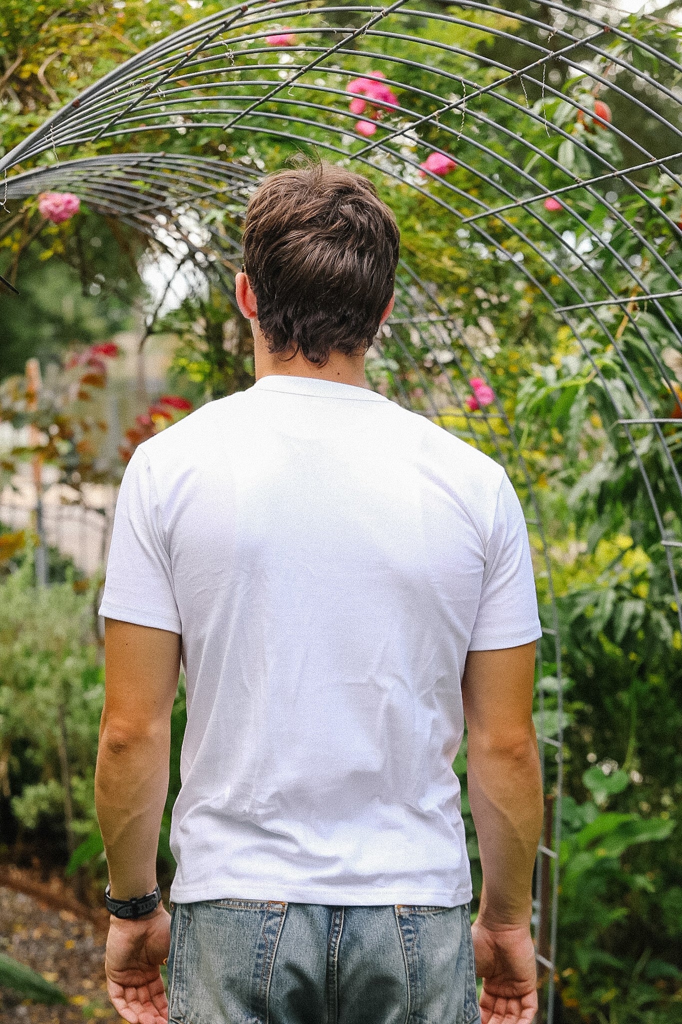 Man wearing a white t-shirt and blue jeans standing under a metal archway in a garden.