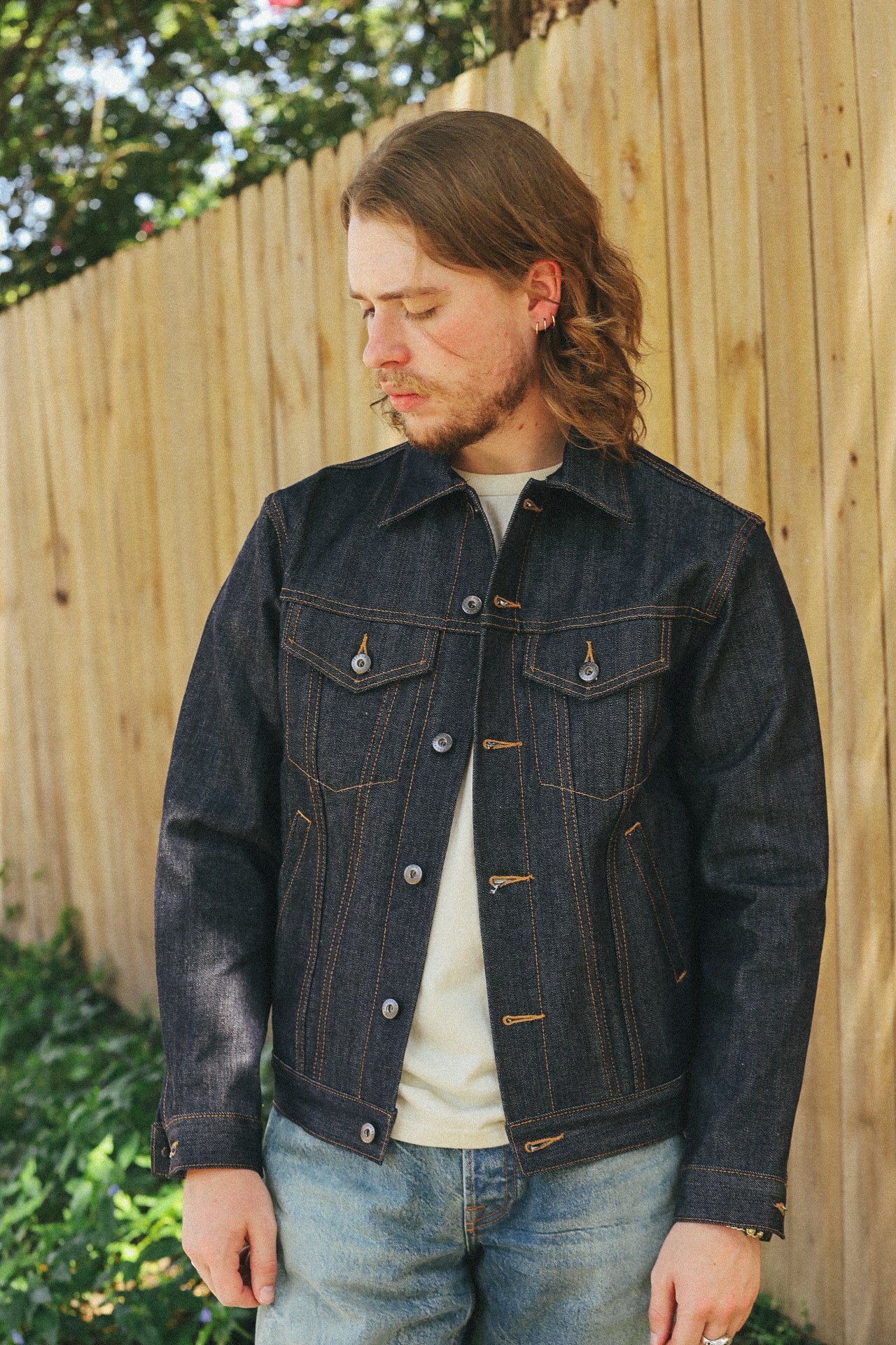 Man wearing dark denim jacket and light jeans standing outdoors by wooden fence with green plants