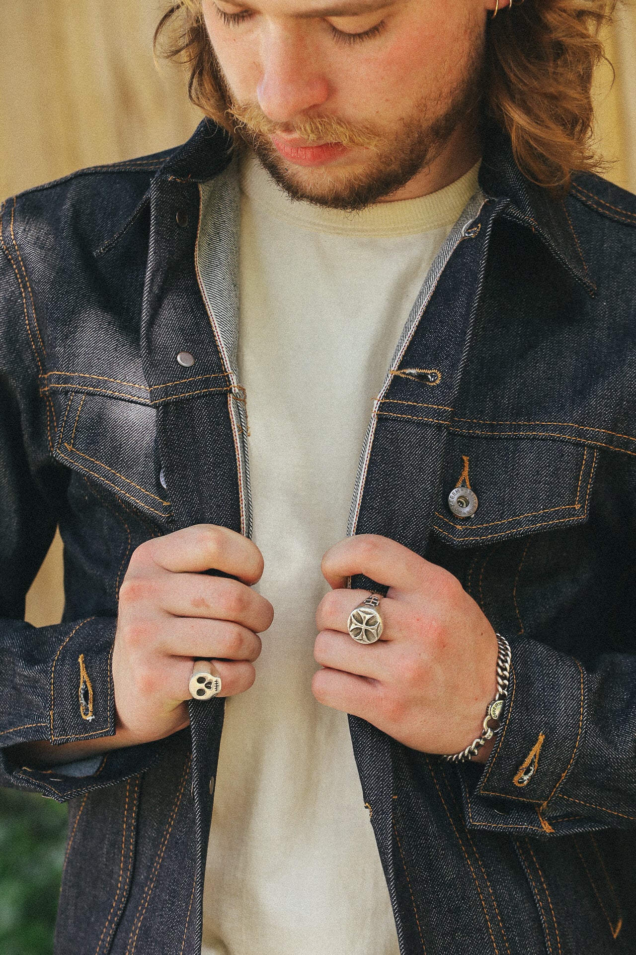 Man wearing dark denim jacket over white shirt, showcasing silver skull and cross rings and chain bracelet