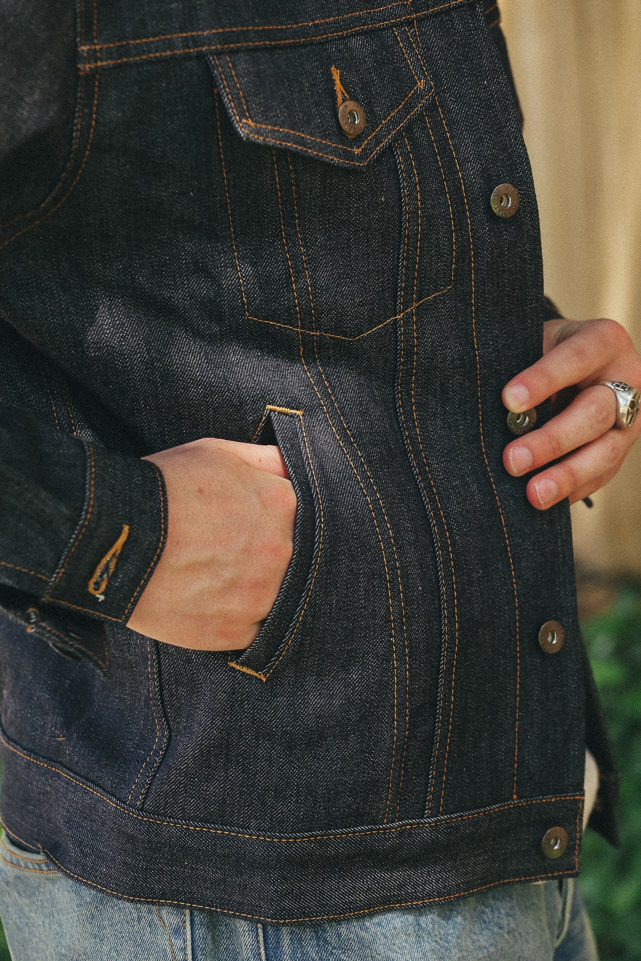 Close-up of person wearing dark denim jacket with orange stitching and hand in pocket