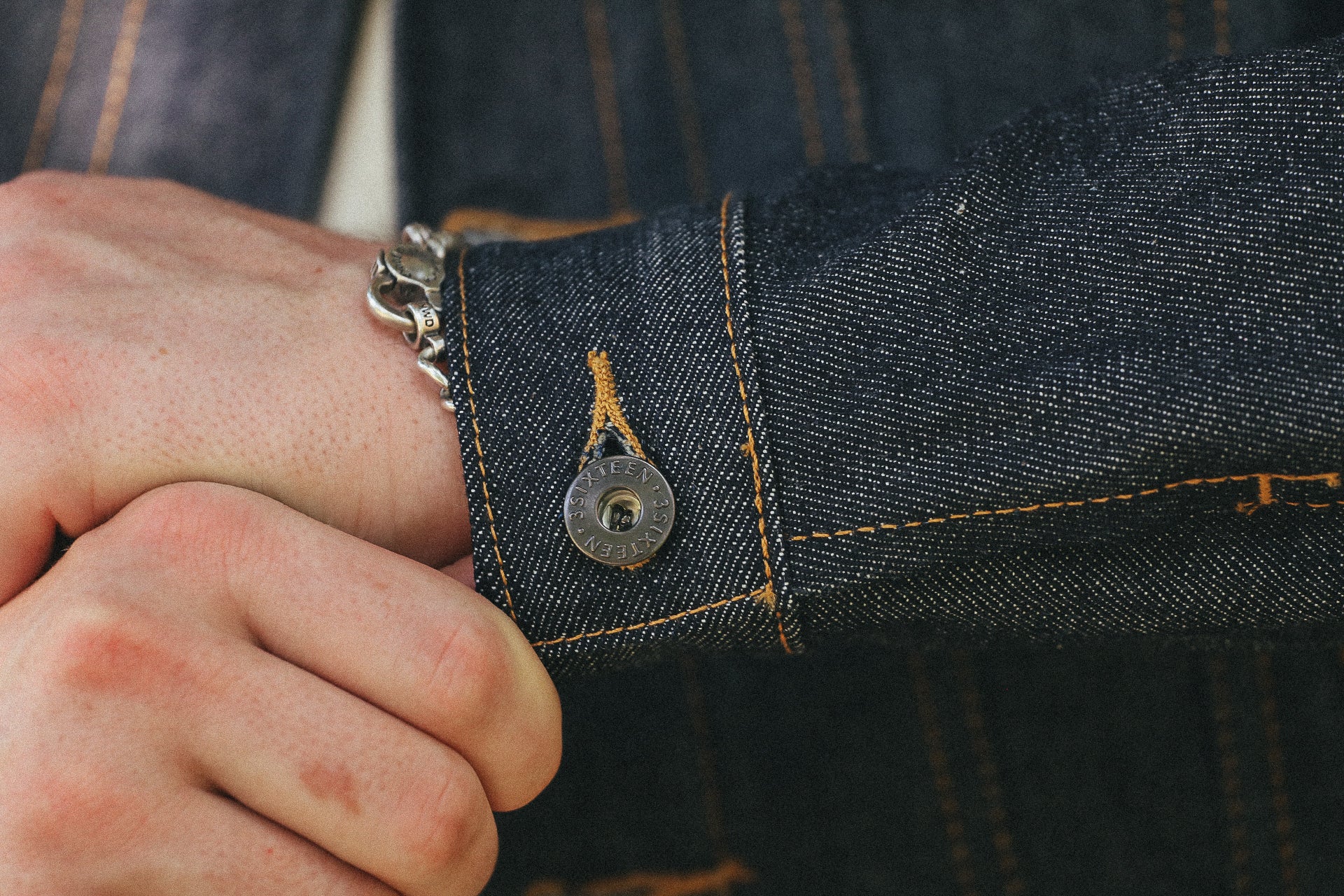 Close-up of hand wearing silver chain bracelet and dark denim jacket cuff with metal button at Iron Shop Provisions