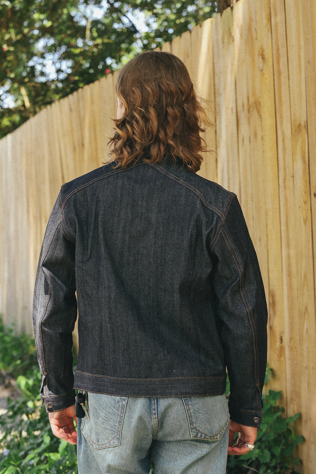 Man with shoulder-length hair wearing dark denim jacket and light blue jeans standing by wooden fence