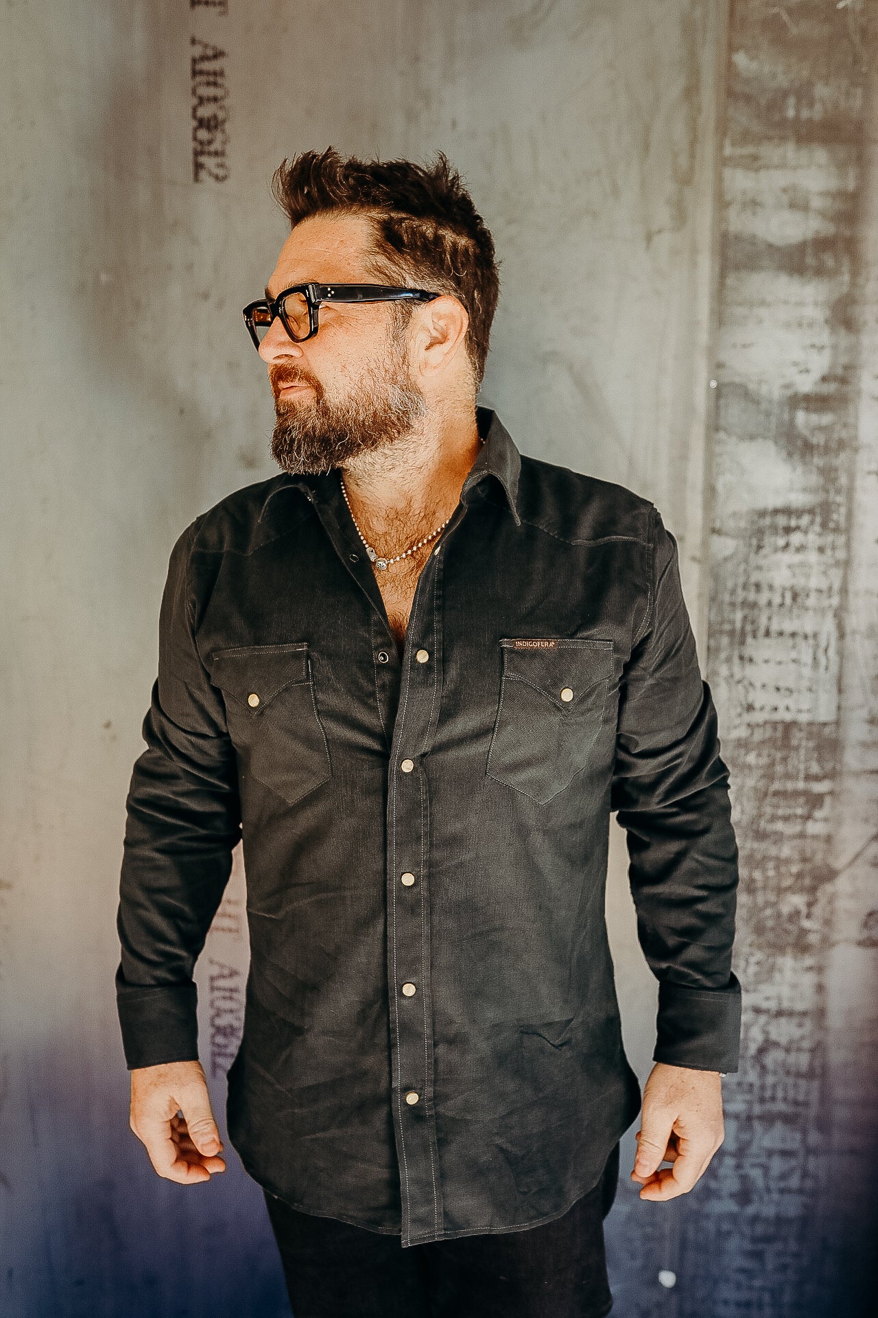 Bearded man wearing black button-up shirt with front pockets and glasses against textured wall