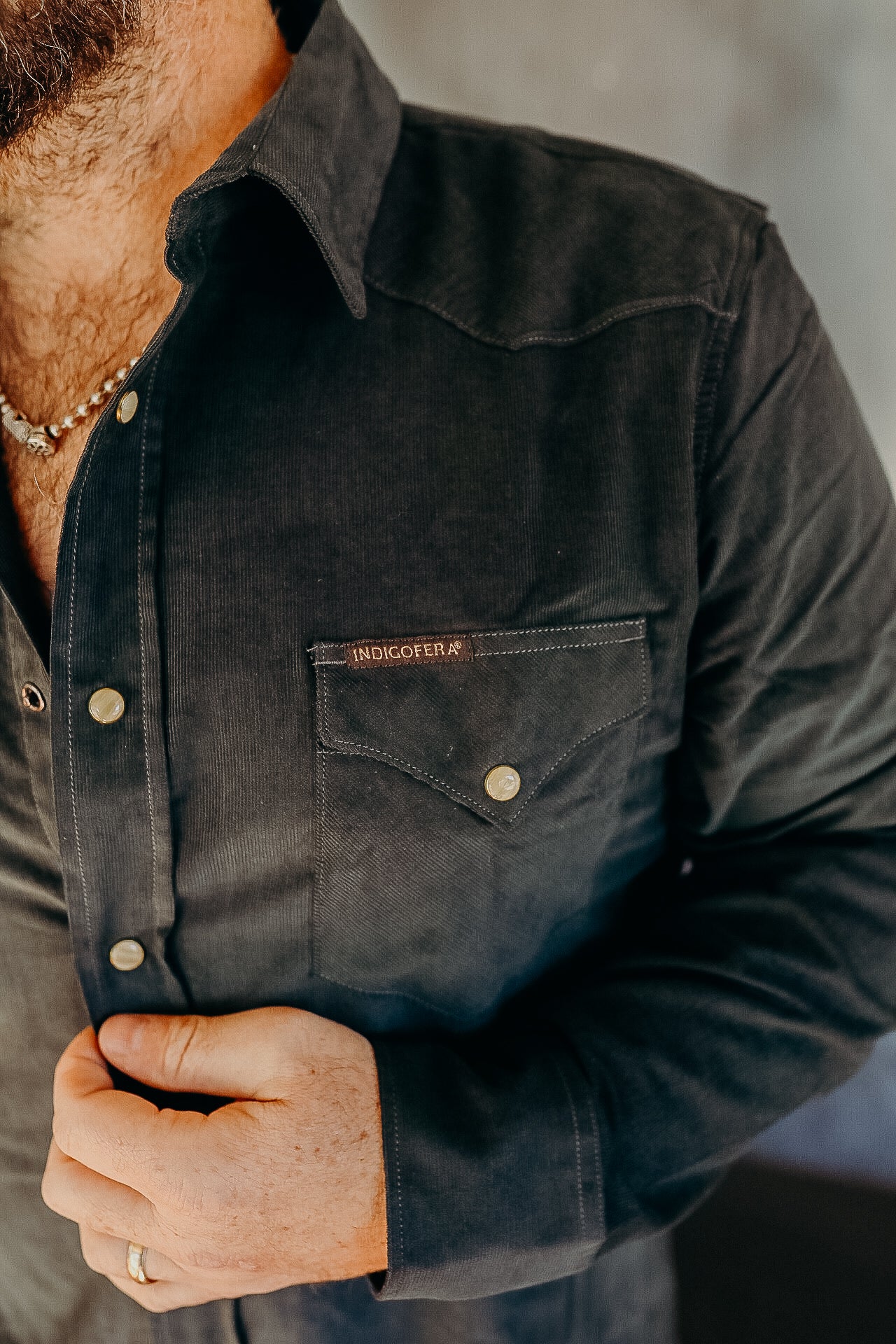 Close-up of a person buttoning a dark Indigofera corduroy shirt with a chest pocket and brass snap buttons