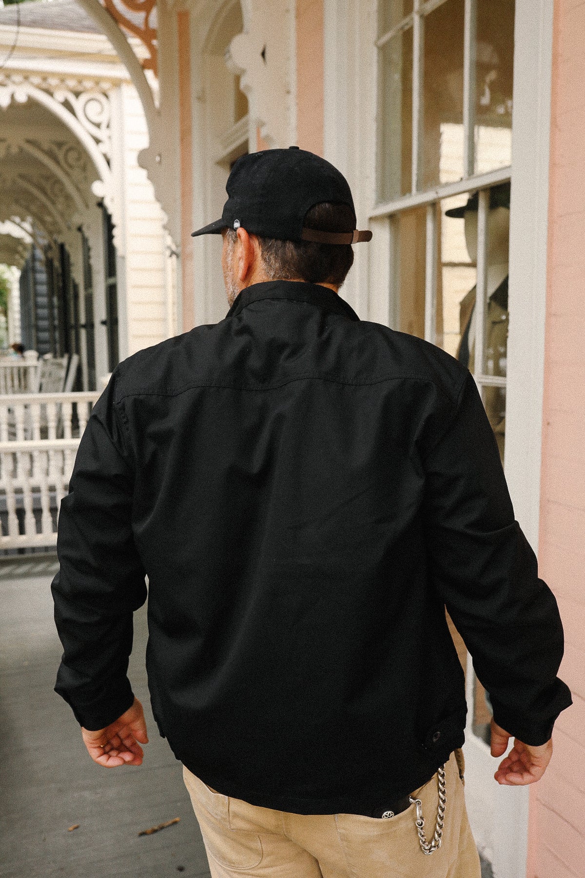 Person wearing a black jacket and cap walking away from a building with decorative architecture.