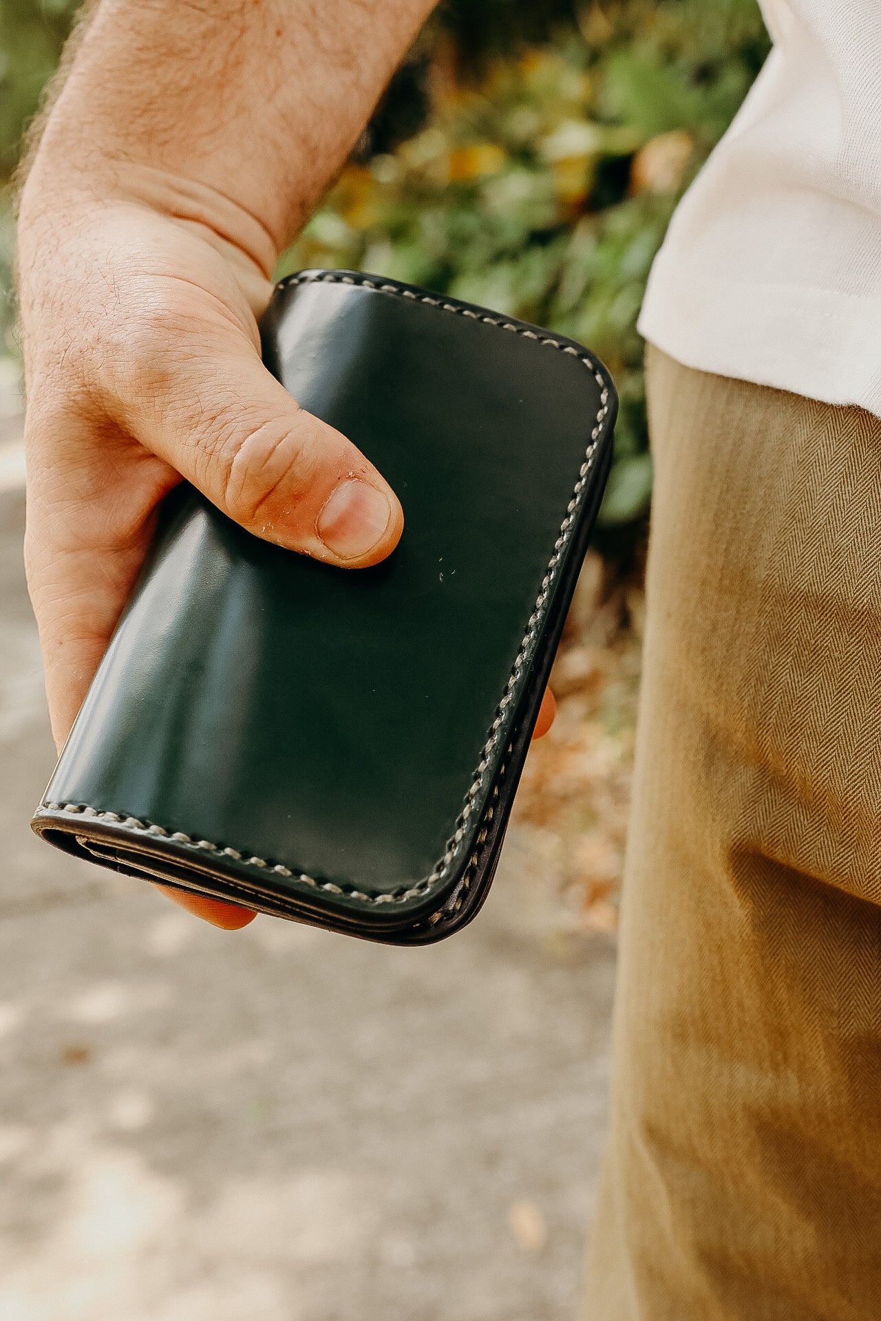 Hand holding black leather wallet with white stitching, wearing tan pants outdoors