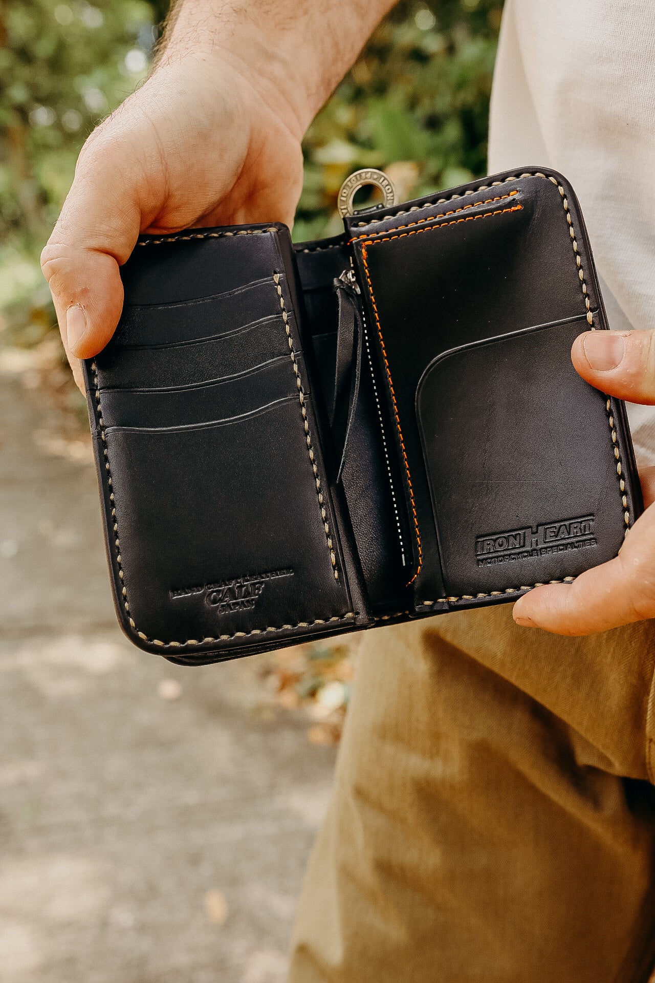 Hands holding an open black leather wallet with card slots and zipper pocket, embossed IRON HEART logo