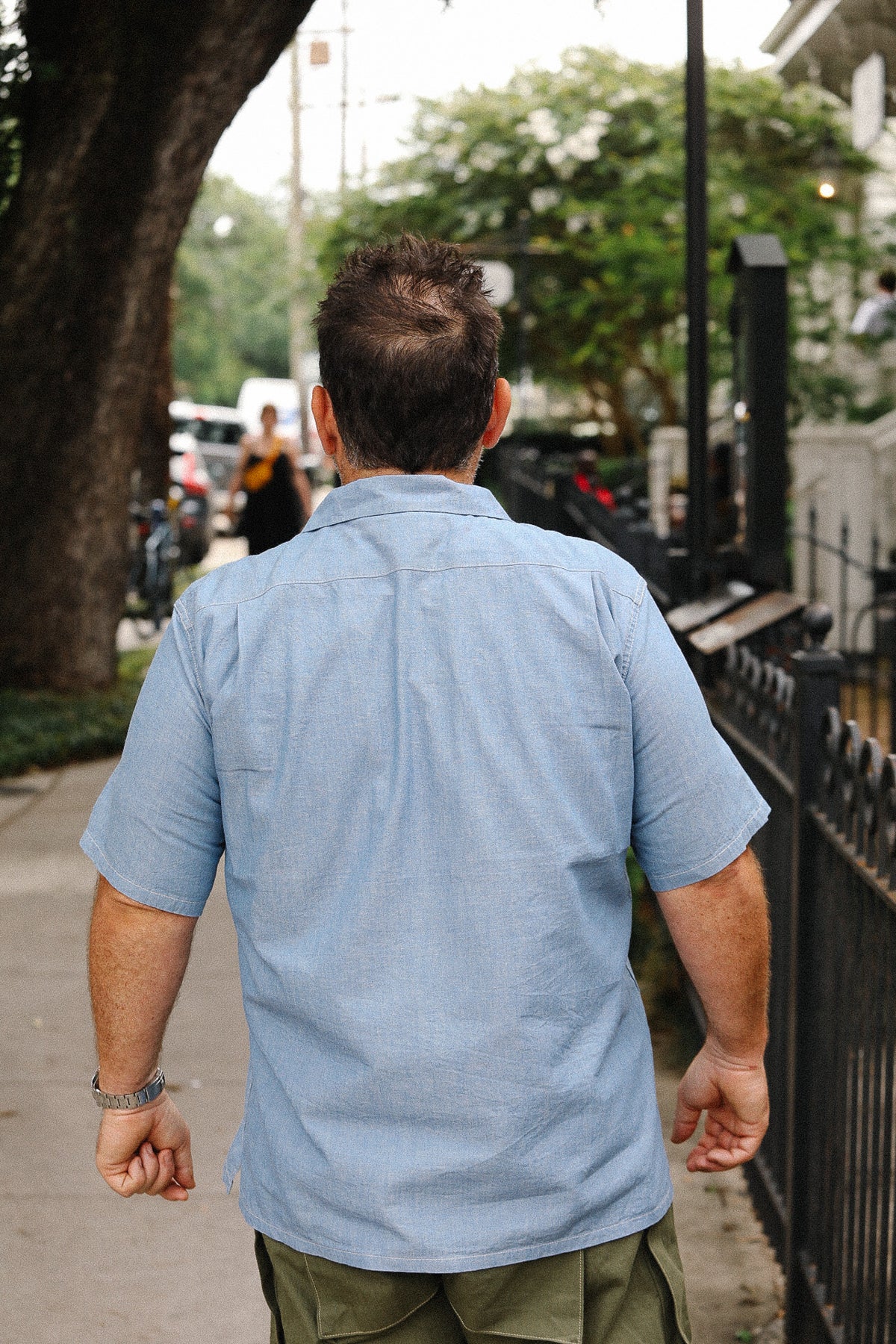 Man in a blue shirt walking on a sidewalk with trees and parked cars in the background