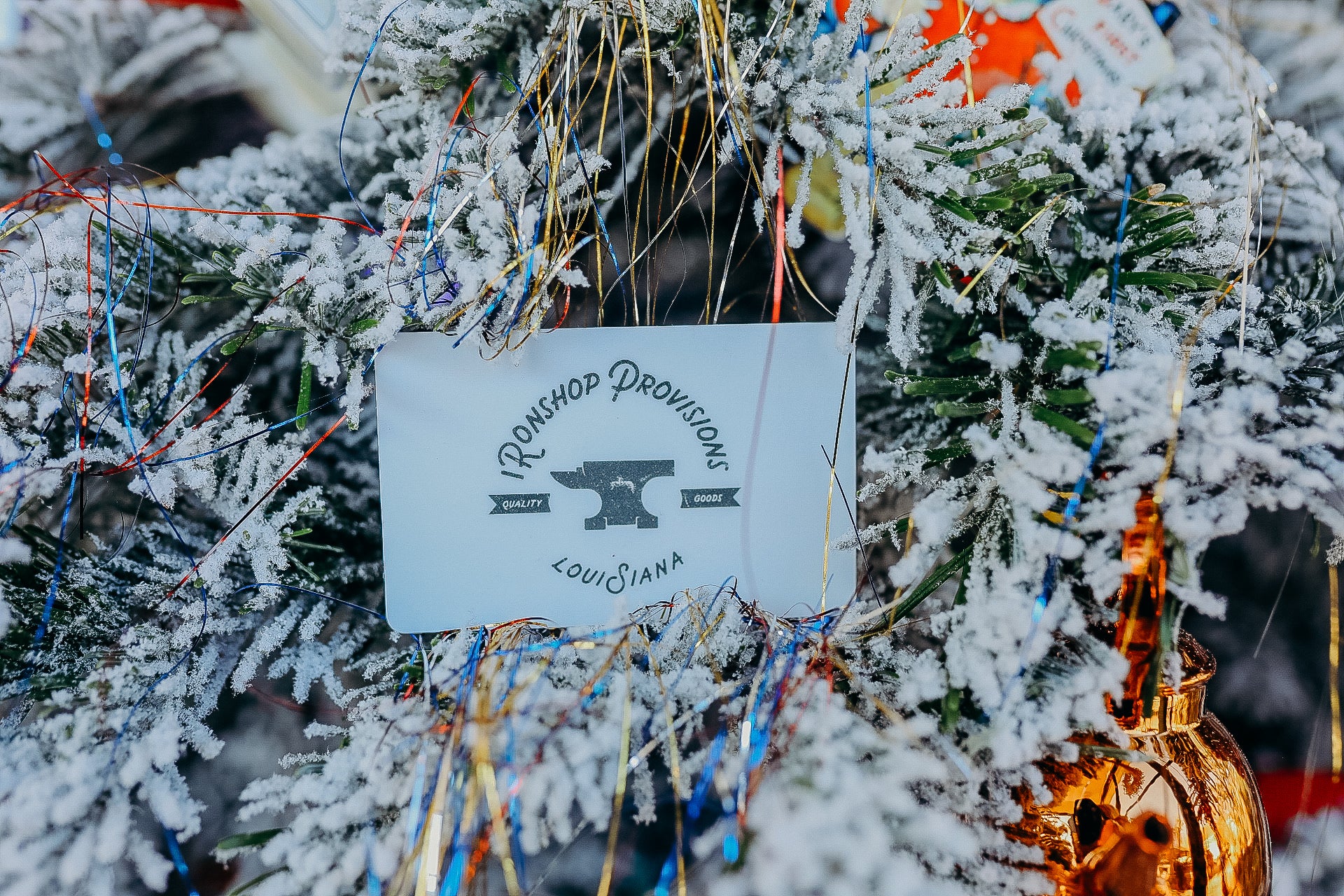 Iron Shop Provisions tag with anvil logo on snow-covered pine branches and colorful tinsel