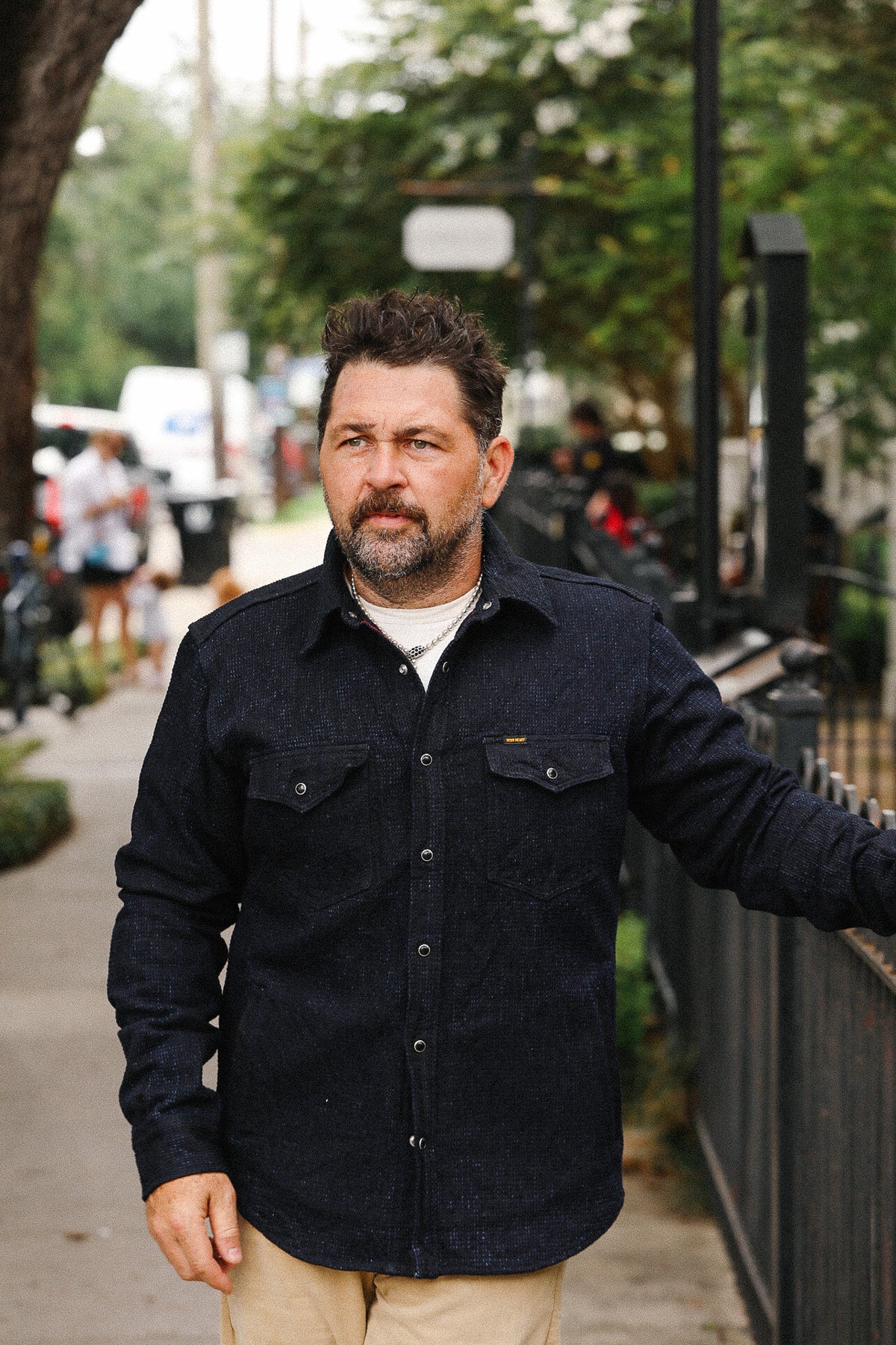 Man wearing a dark jacket standing on a sidewalk with trees and people in the background