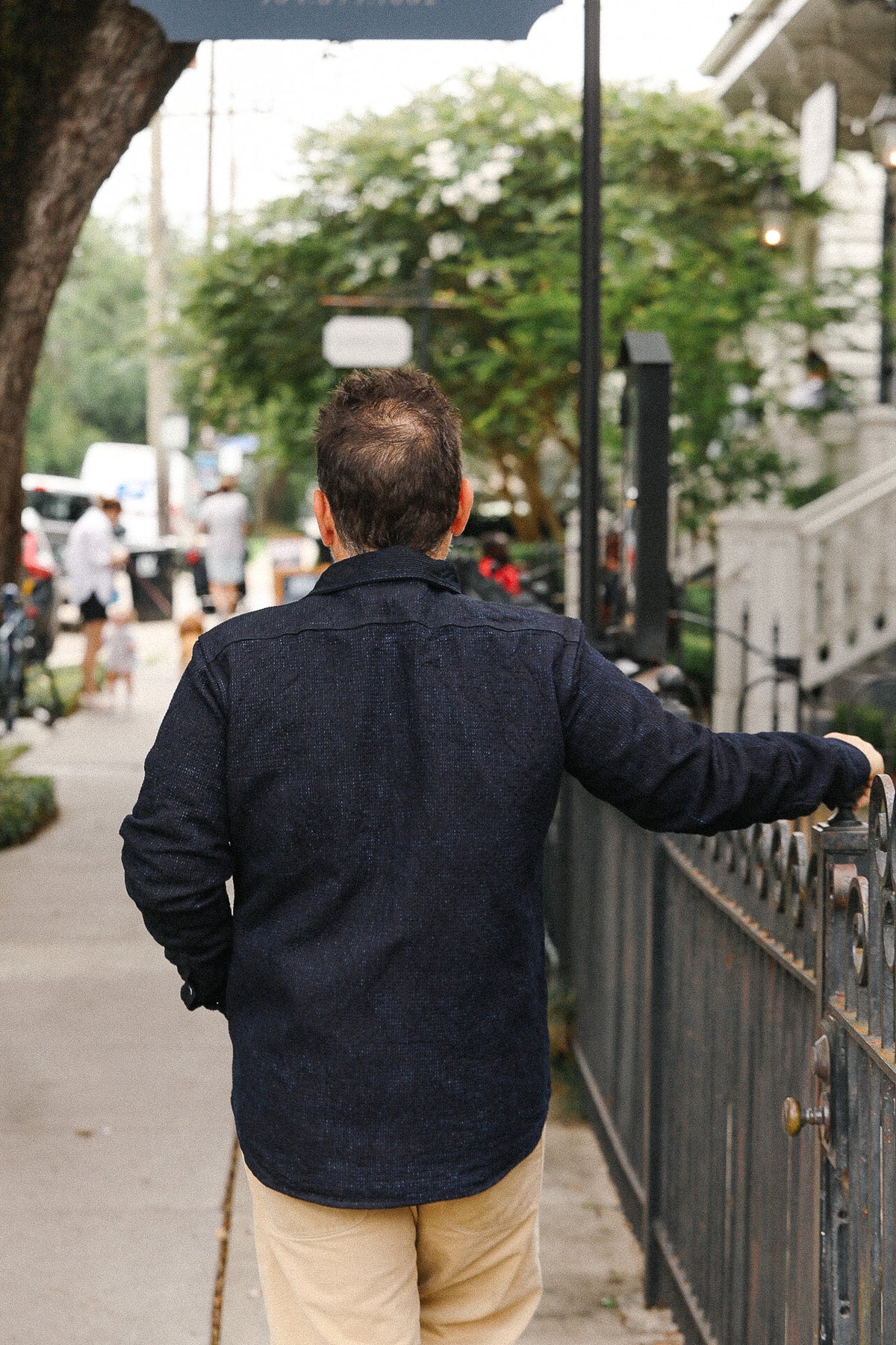 Man in a dark jacket walking on a sidewalk with trees and people in the background