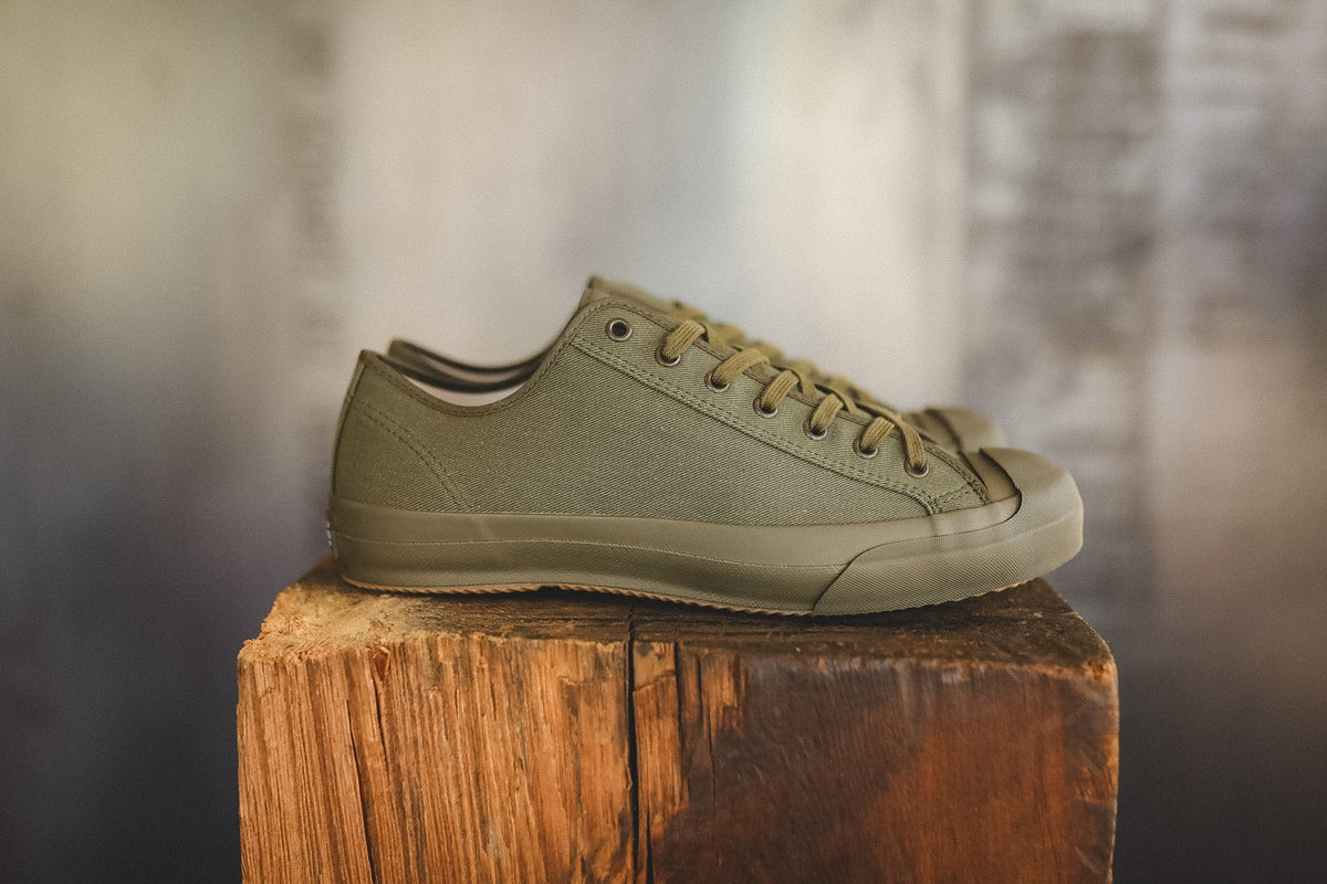 Up close image of a olive green sneaker on a wood block with a grey background 