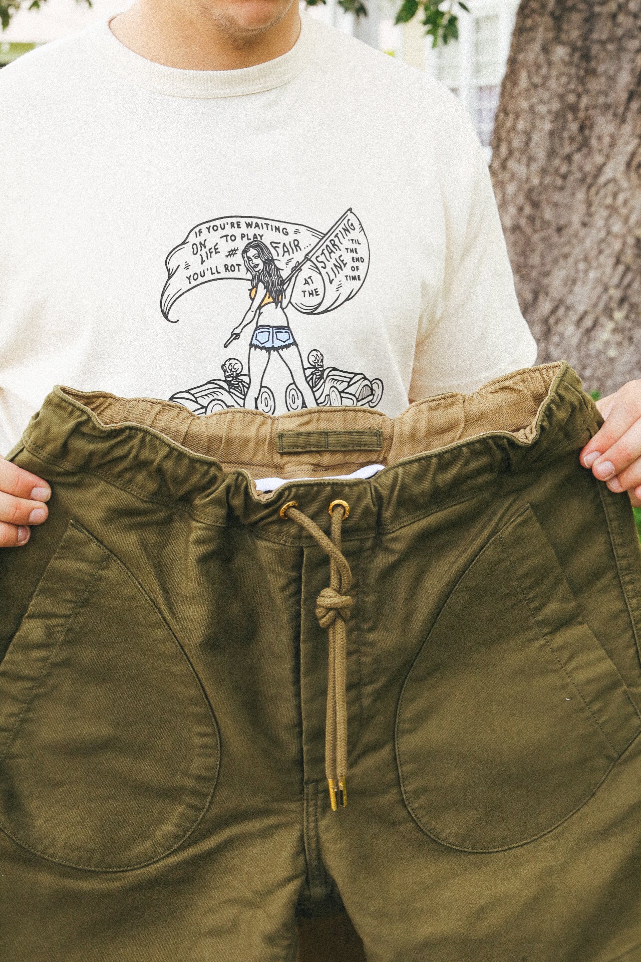Person holding olive green drawstring pants in front of a white graphic t-shirt outdoors