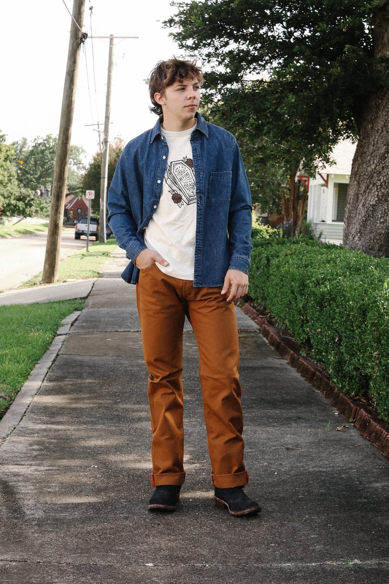 Man wearing casual outfit with denim shirt, graphic t-shirt, brown pants, and black boots on sidewalk
