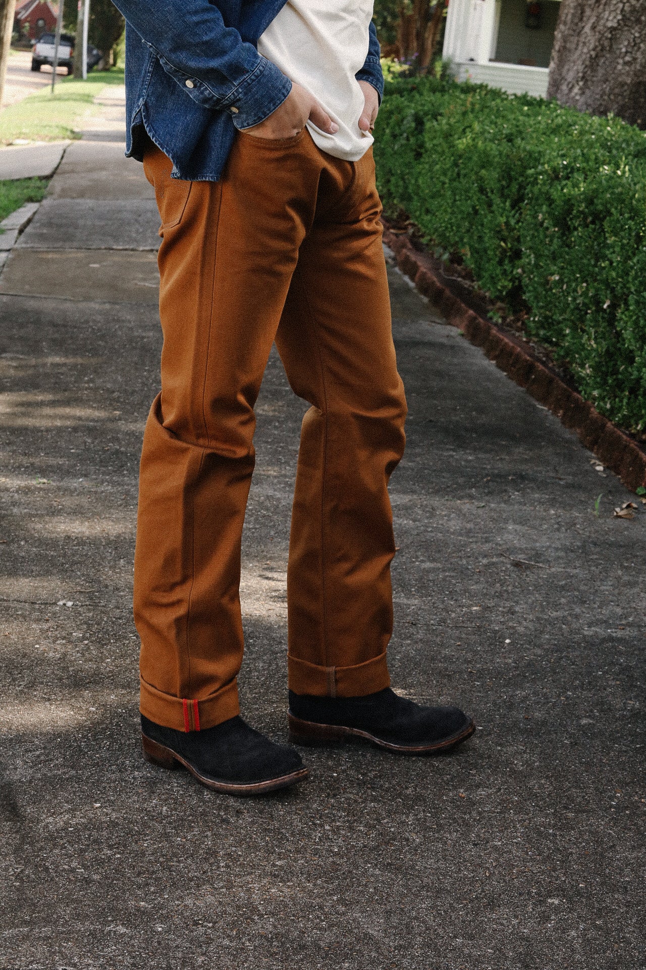 Person wearing brown work pants, black boots, white shirt, and a denim jacket outdoors on sidewalk