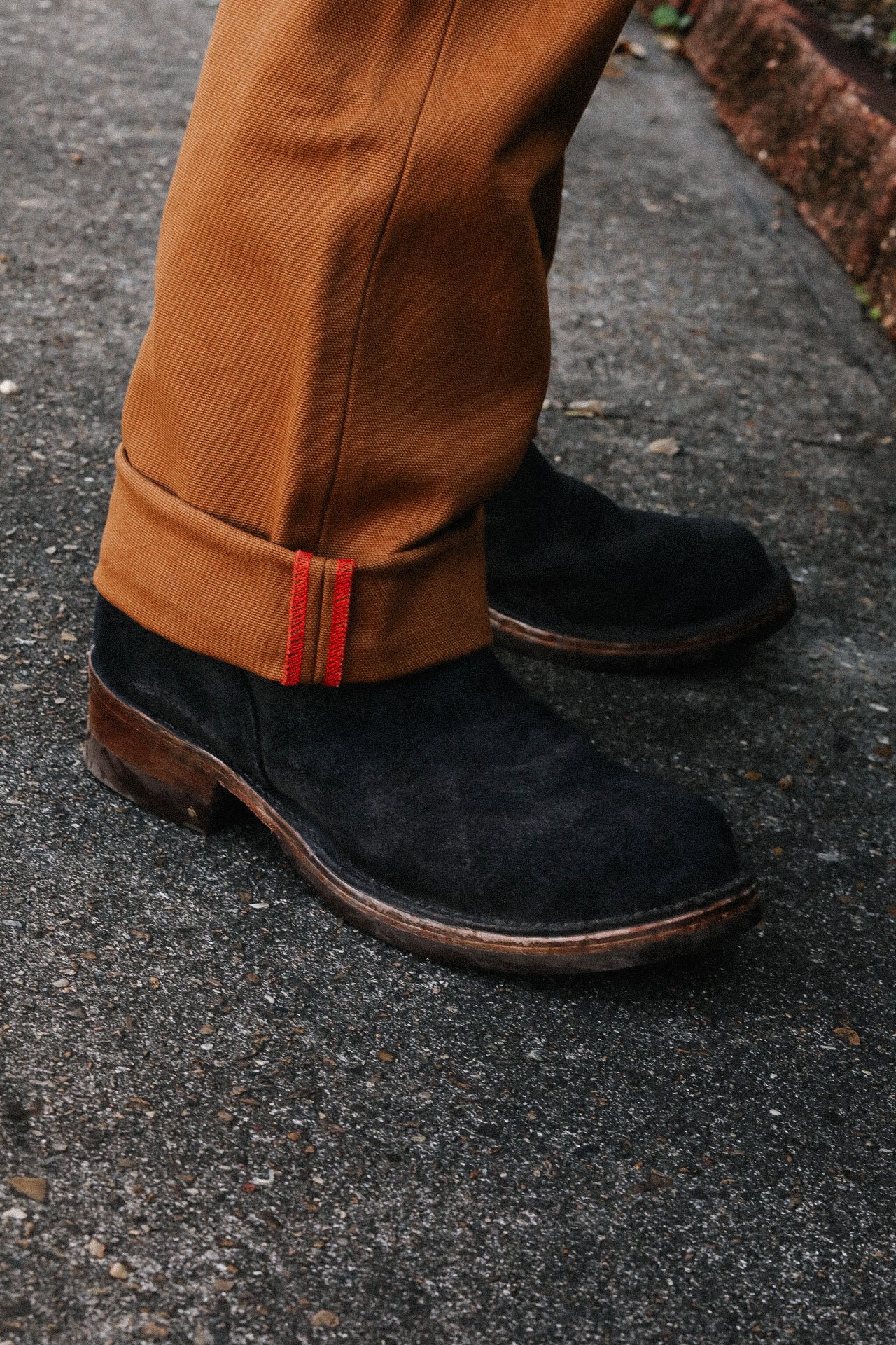 Close-up of black suede boots and rolled-up brown pants with red stitching detail on asphalt