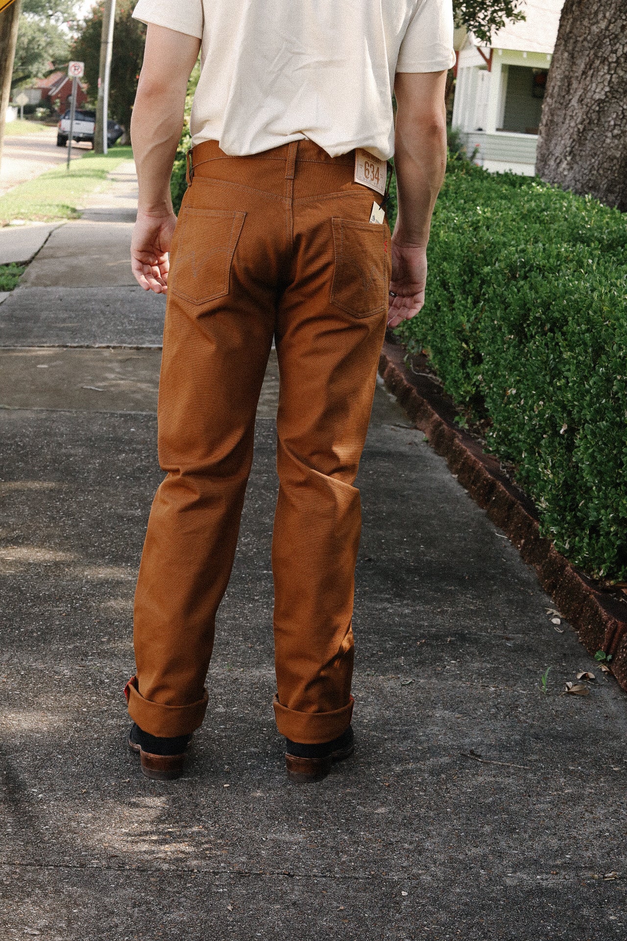 Man wearing brown work pants standing on a sidewalk near green hedges in a suburban neighborhood