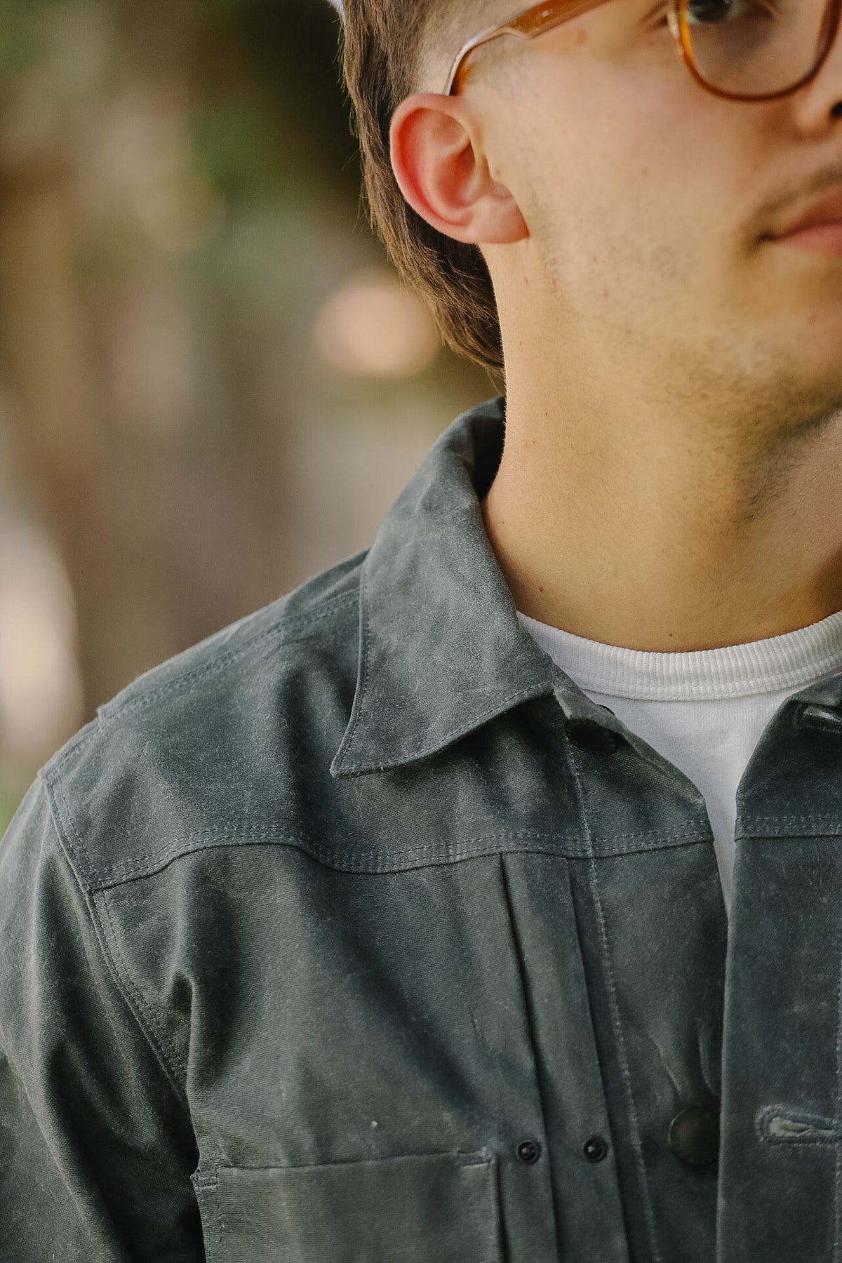 Close-up of a man wearing a dark charcoal gray canvas utility jacket over a white t-shirt, with brown-rimmed glasses, set against a soft, blurred outdoor background.