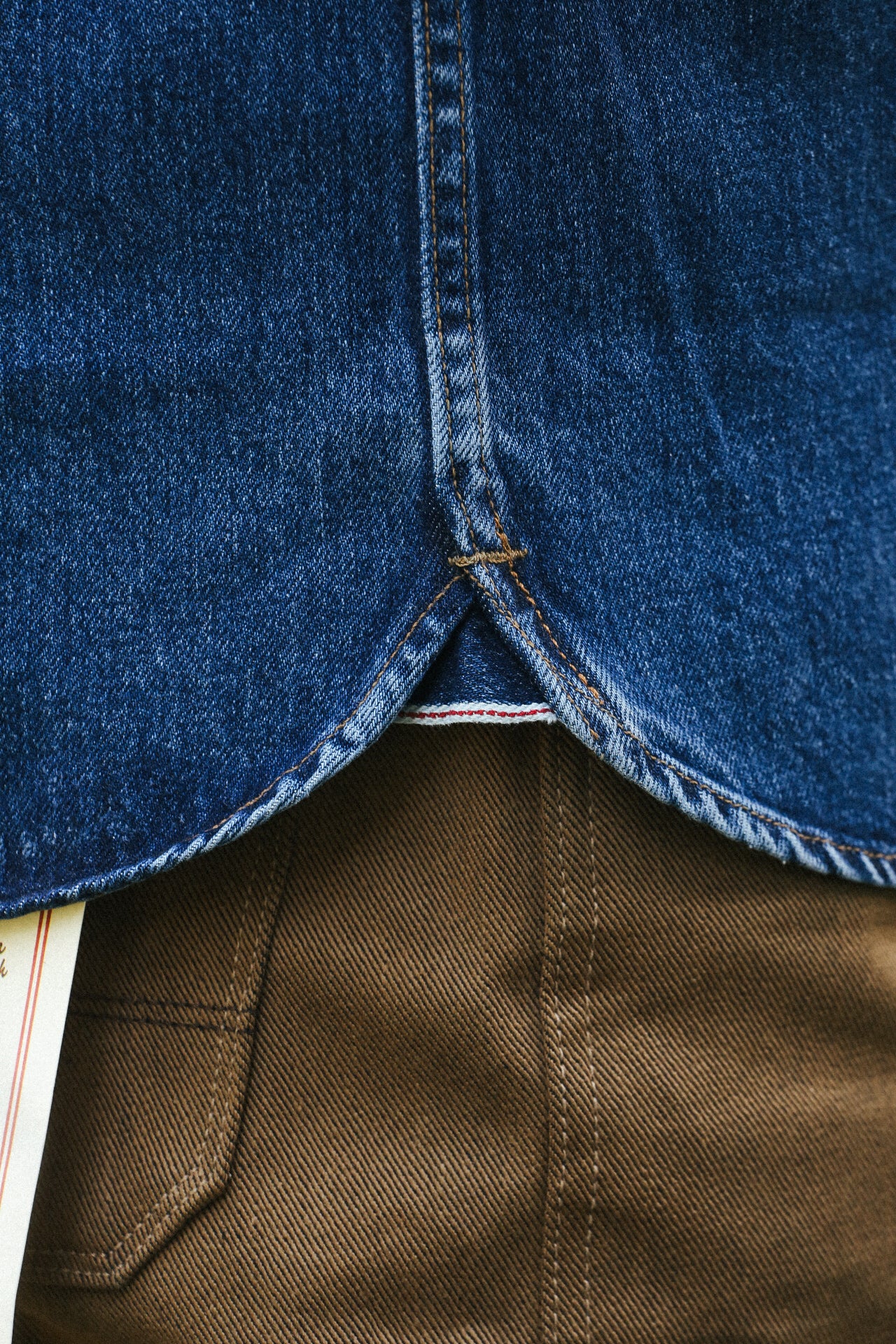 Close-up of blue denim shirt hem with brown workwear pants pocket, detailed stitching visible