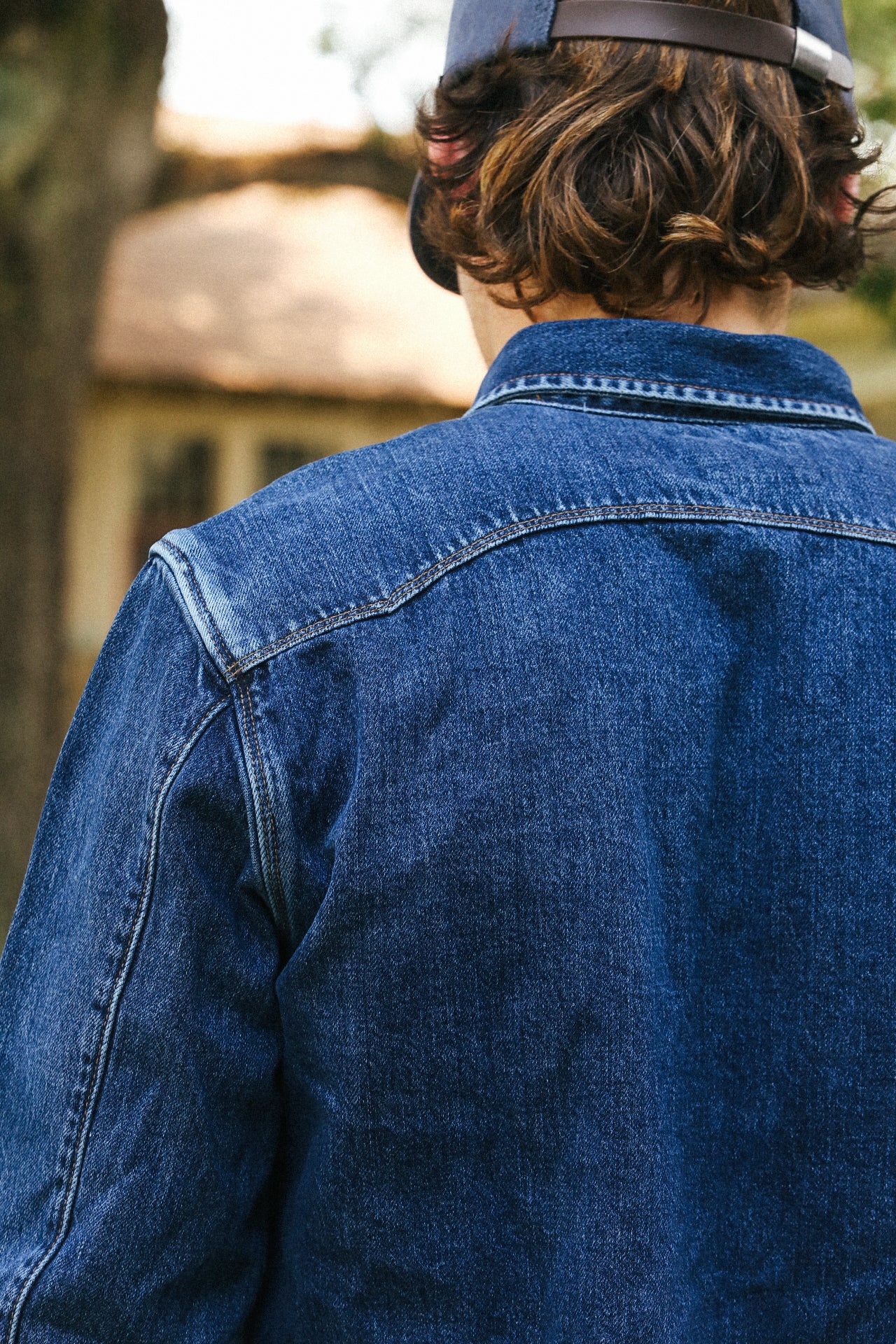 Back view of person wearing blue denim jacket and navy cap outdoors in soft focus background