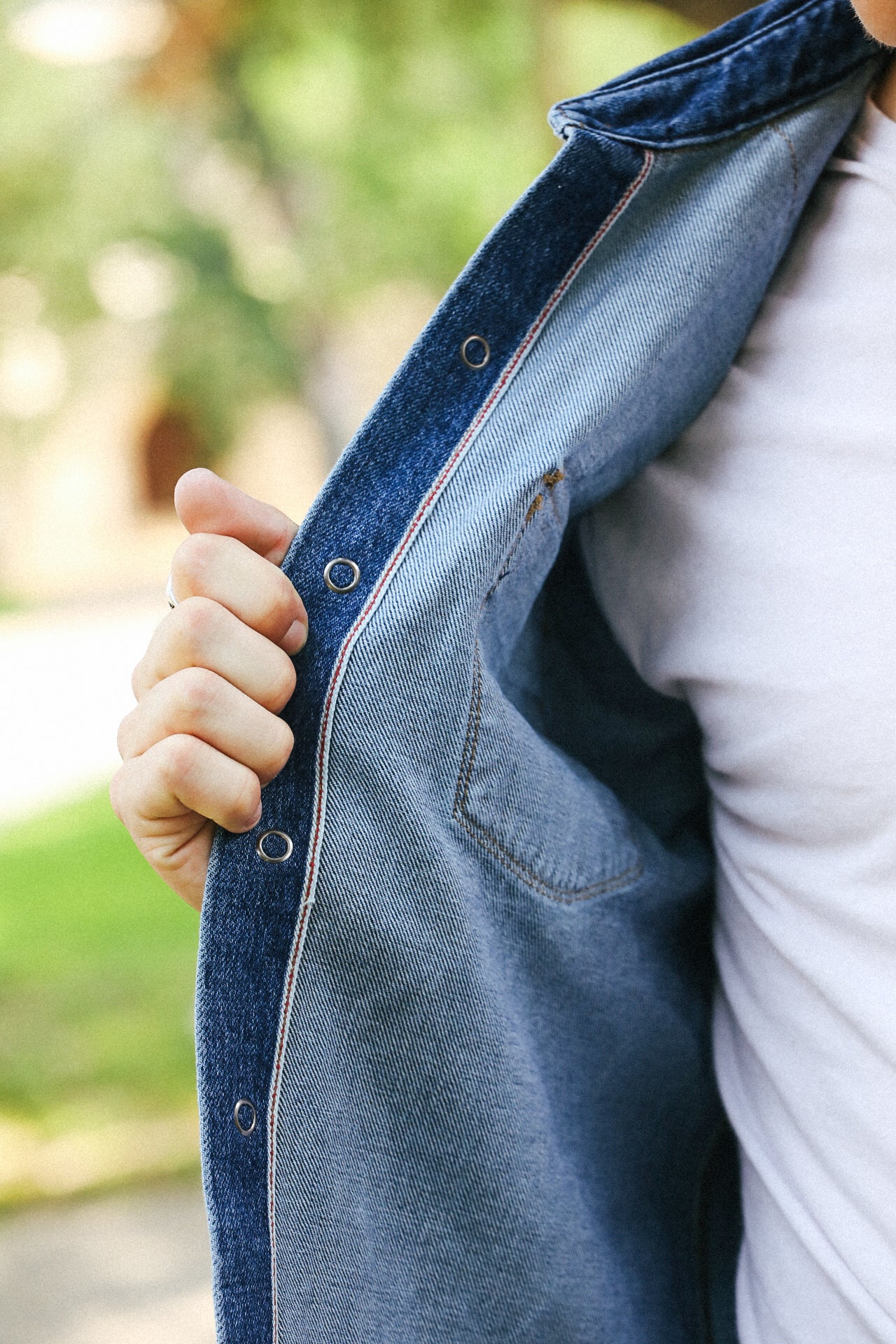 Person holding open the inner side of a blue denim jacket showing stitching and a pocket, wearing a white shirt