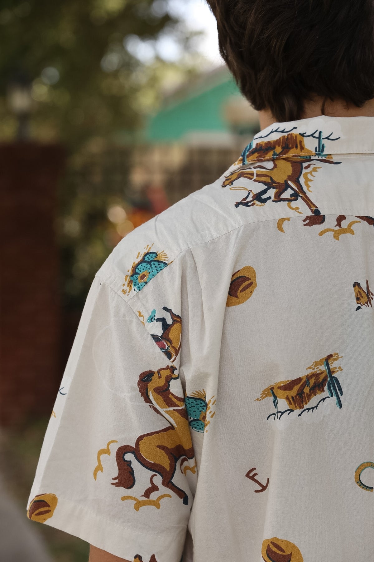 A close-up back view of a person wearing a white short-sleeve button-up shirt with a colorful printed pattern of cowboy horses, hats, and boots, set against a blurred outdoor background.