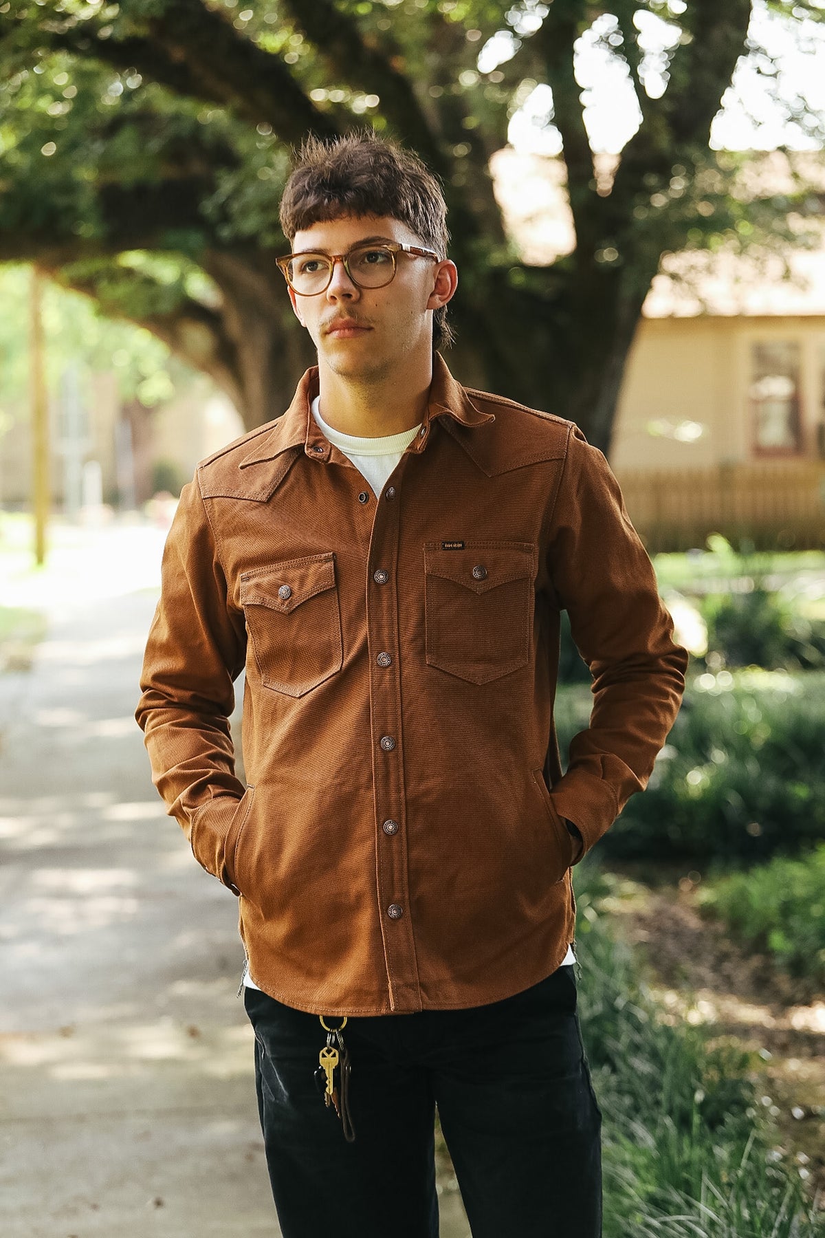 A man wearing a brown corduroy western-style button-up shirt and black pants, standing outdoors on a path with trees in the background.