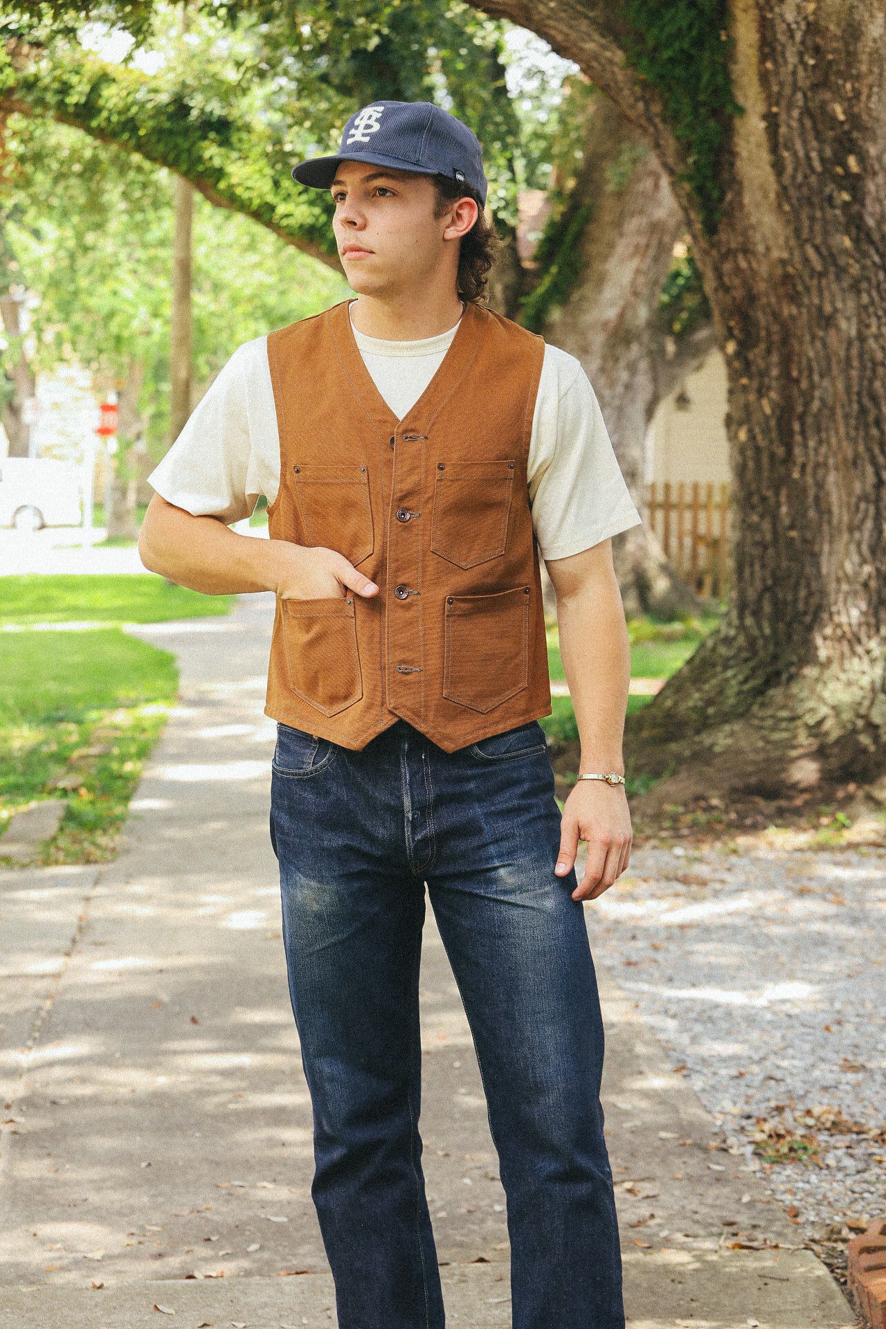 Young man wearing brown button-up work vest and dark denim jeans outdoors on shaded sidewalk