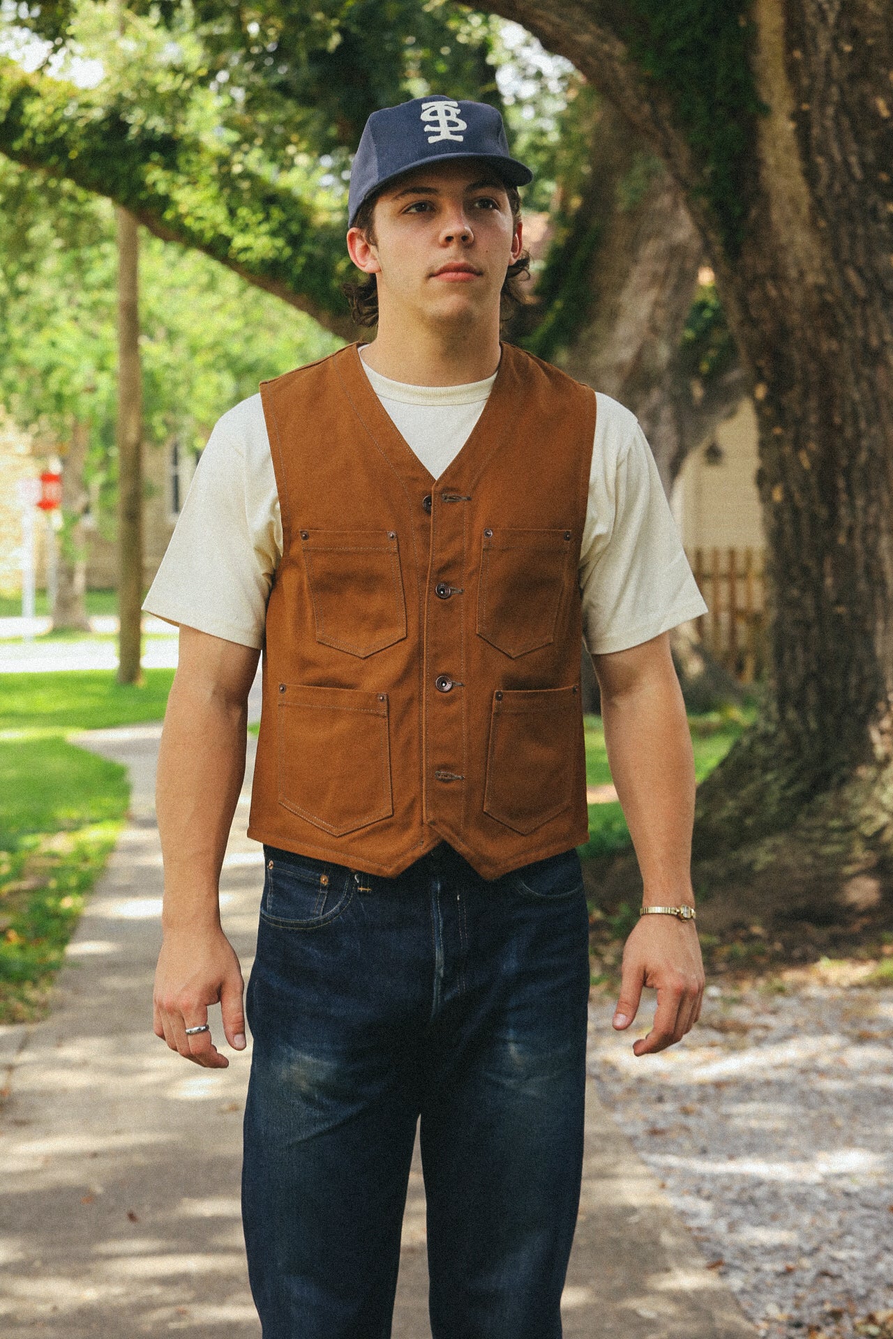 Man wearing a brown workwear vest, white t-shirt, dark jeans, and a navy cap, standing outdoors on a tree-lined path